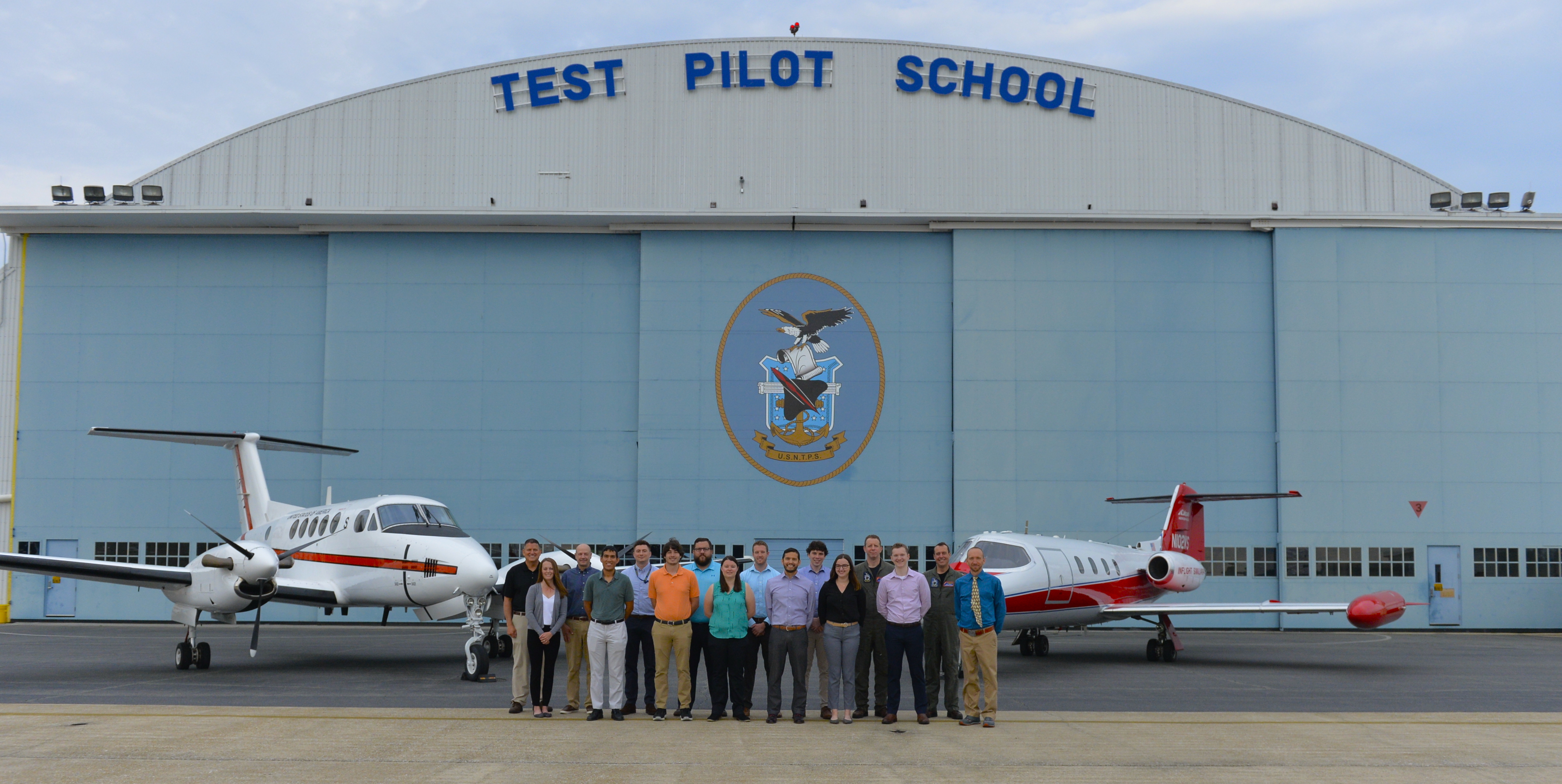 Civilian engineers from NAWCAD line up for a photo in front of U.S. Naval Test Pilot School's hangar in front of schoolhouse aircraft.