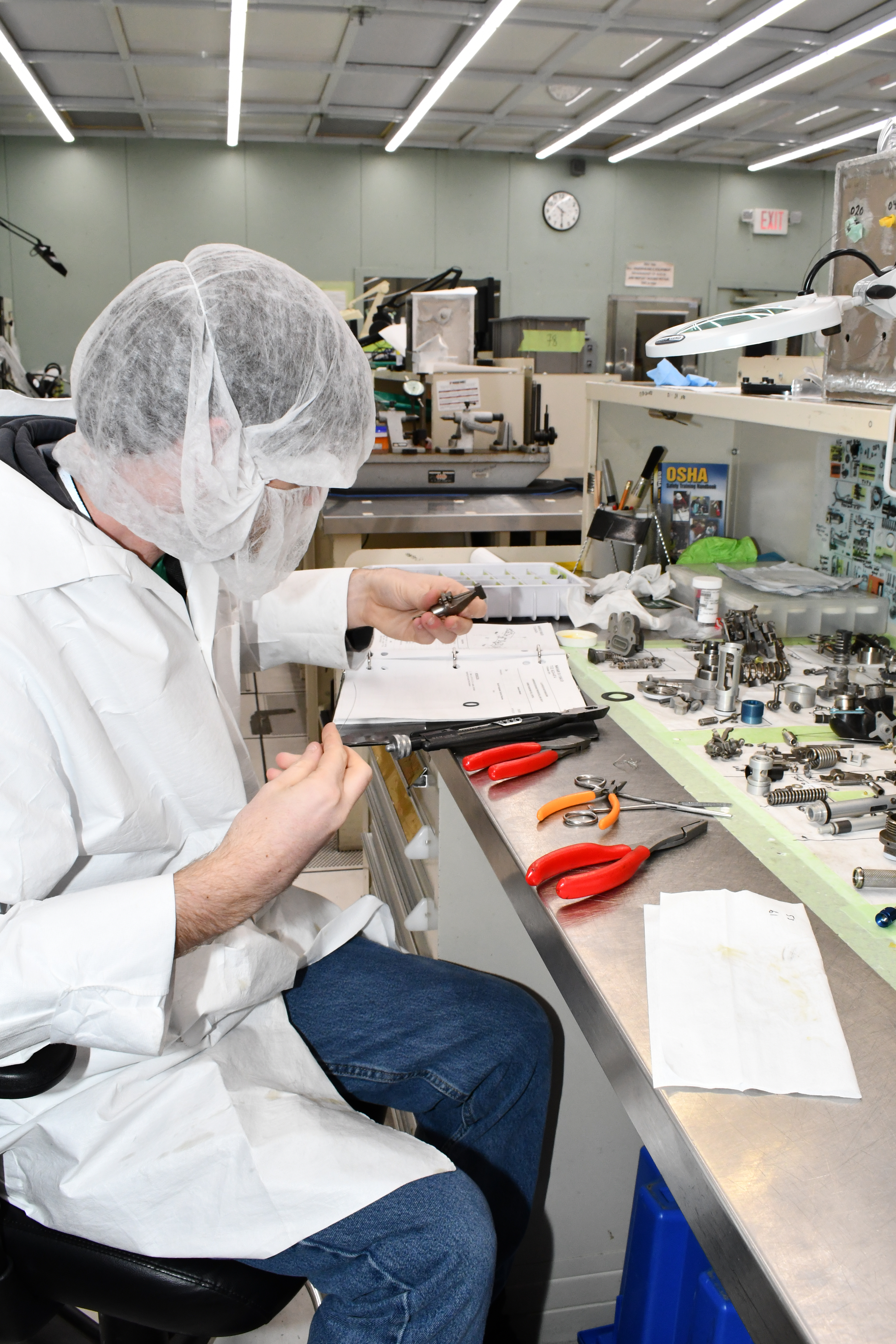 Man in lab coat and hair net and beard cover assembles fuel control unit at clean room work bench
