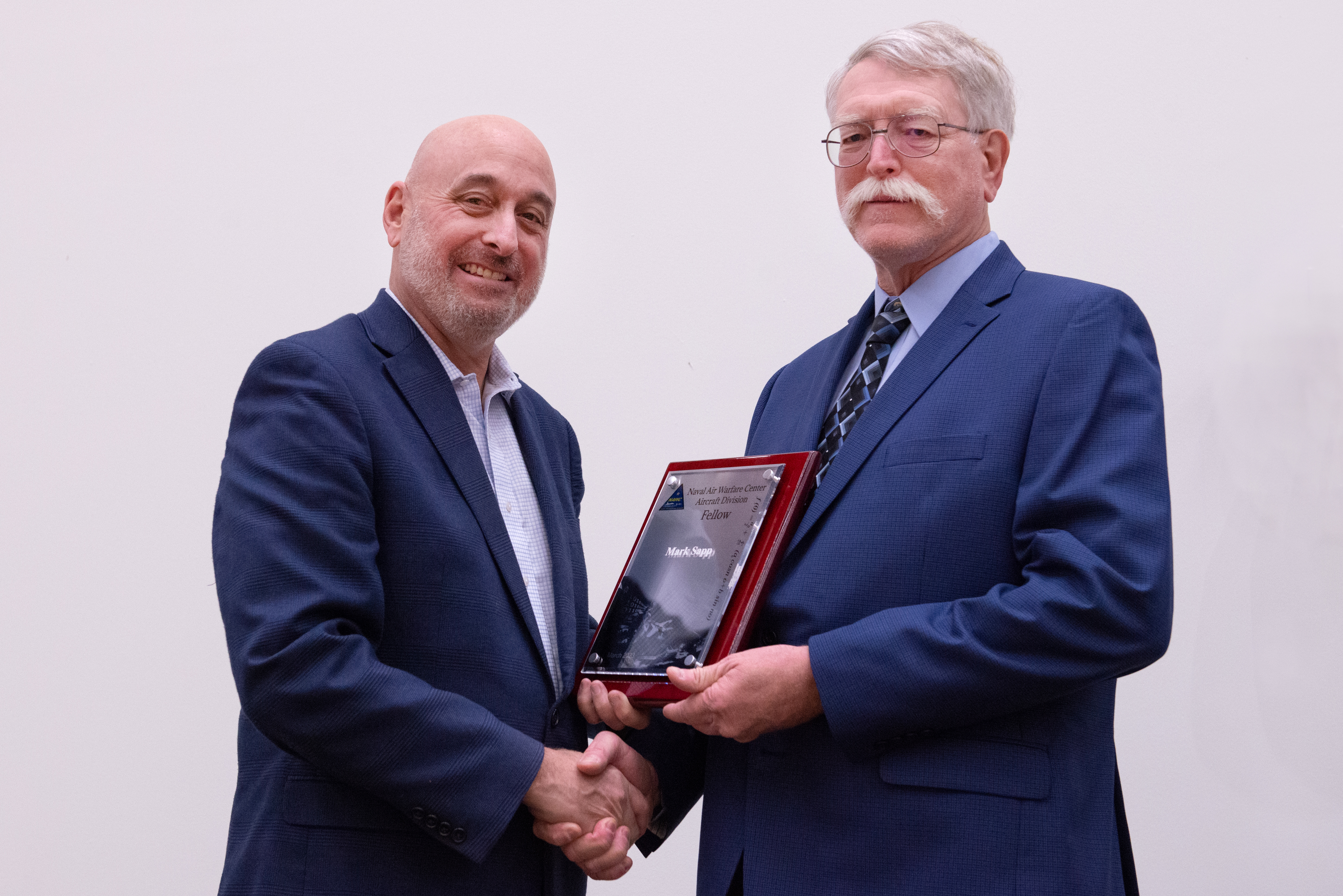 Two men in blue suits shake hands and pose for photo with award plaque between them