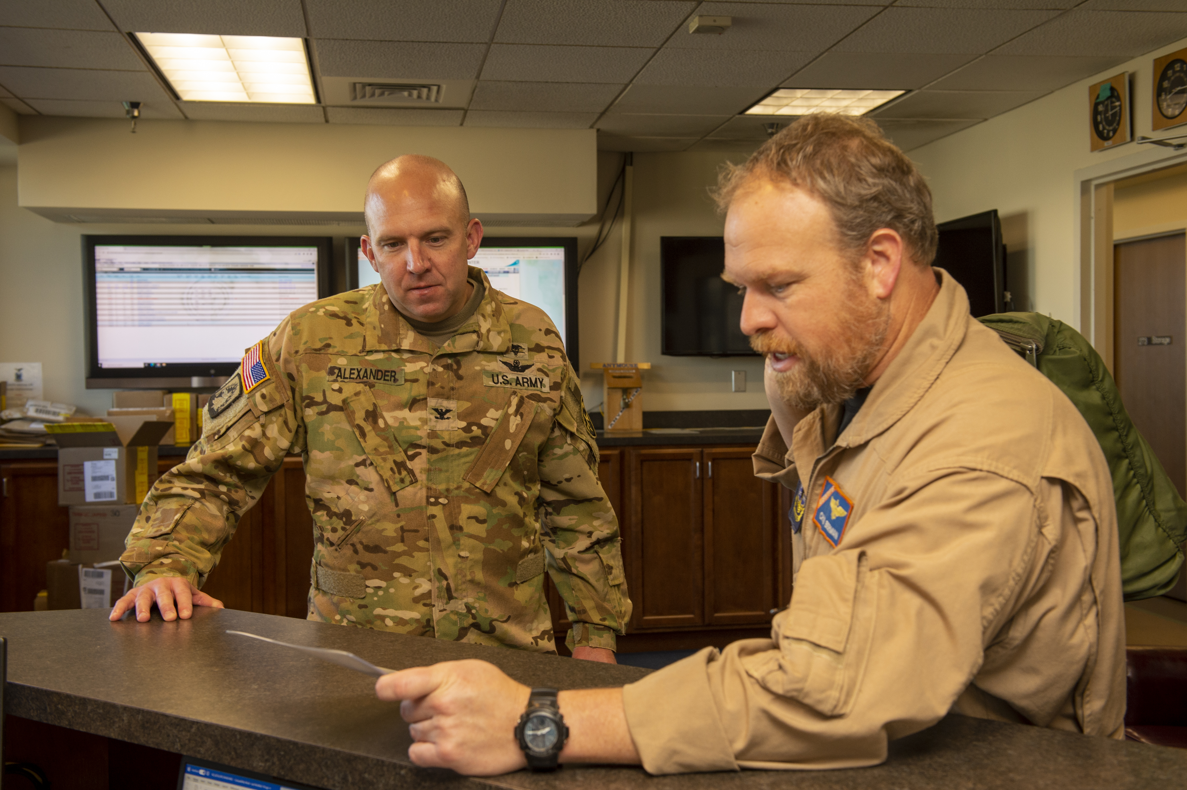 A man in Navy uniform shows a log book to a man in Army uniform in an office space.