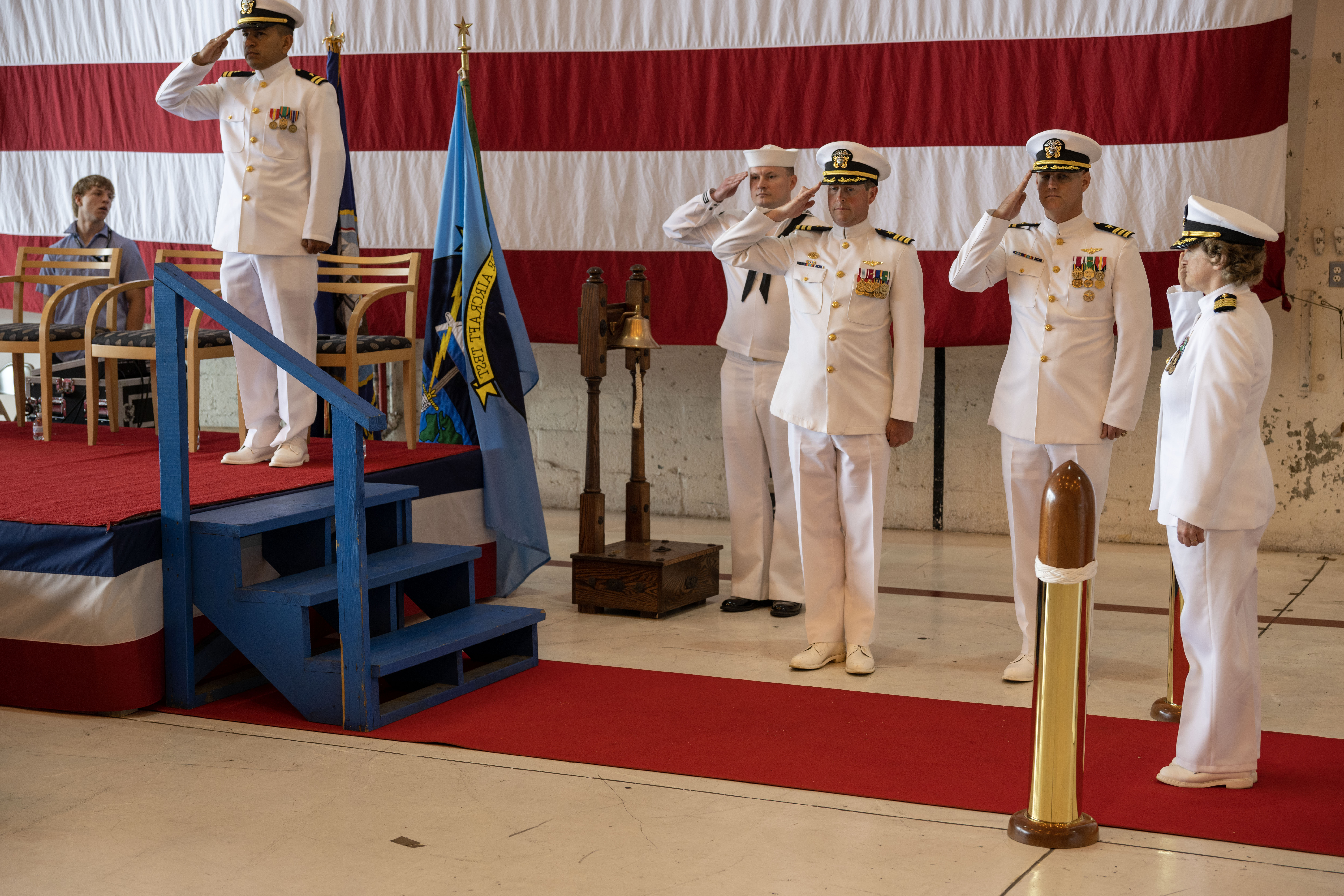 Officers in white Navy dress uniforms salute in front of a ceremonial stage.