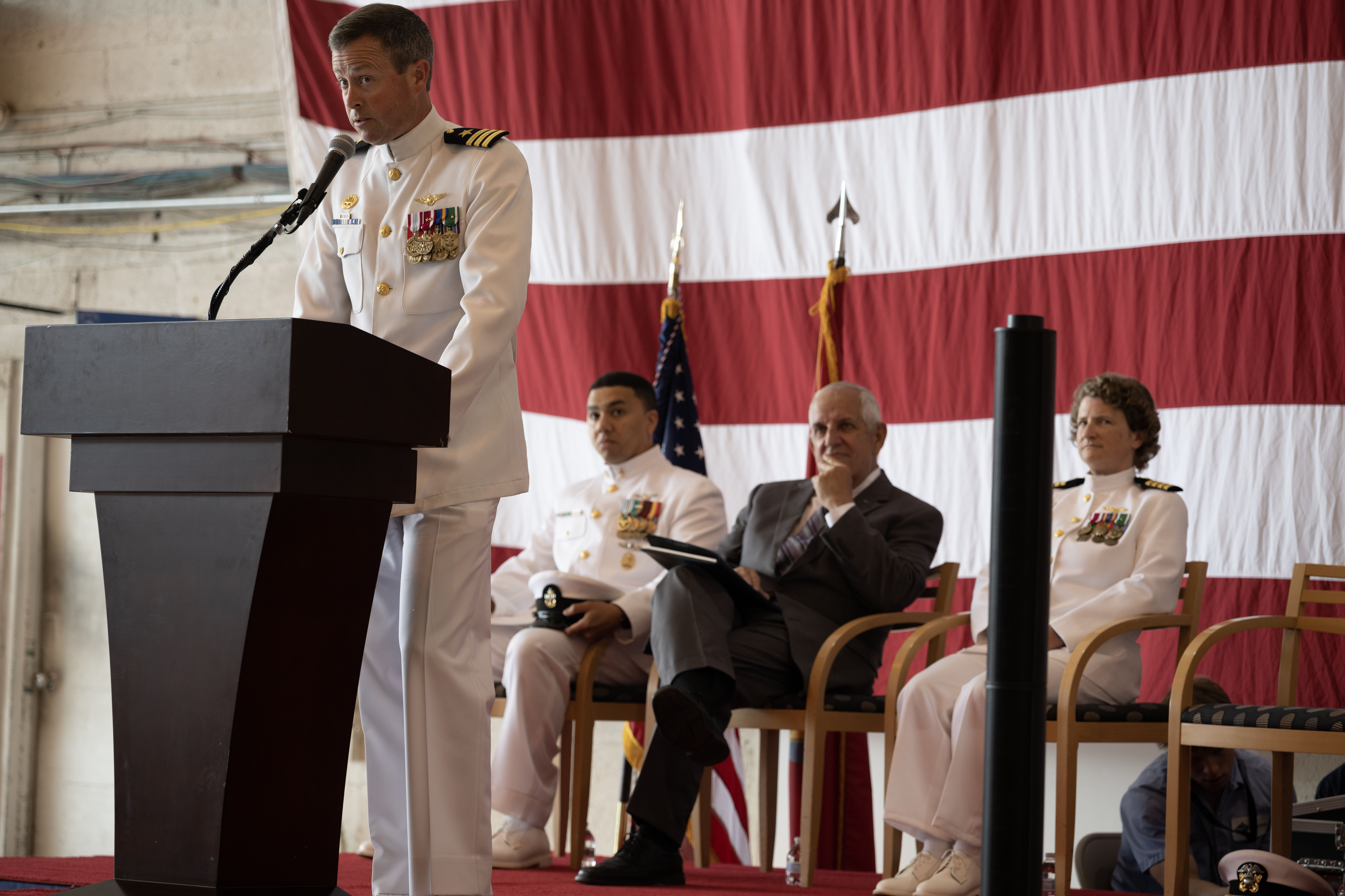 An officer in a white Navy dress uniform speaks behind a podium against an American flag backdrop.