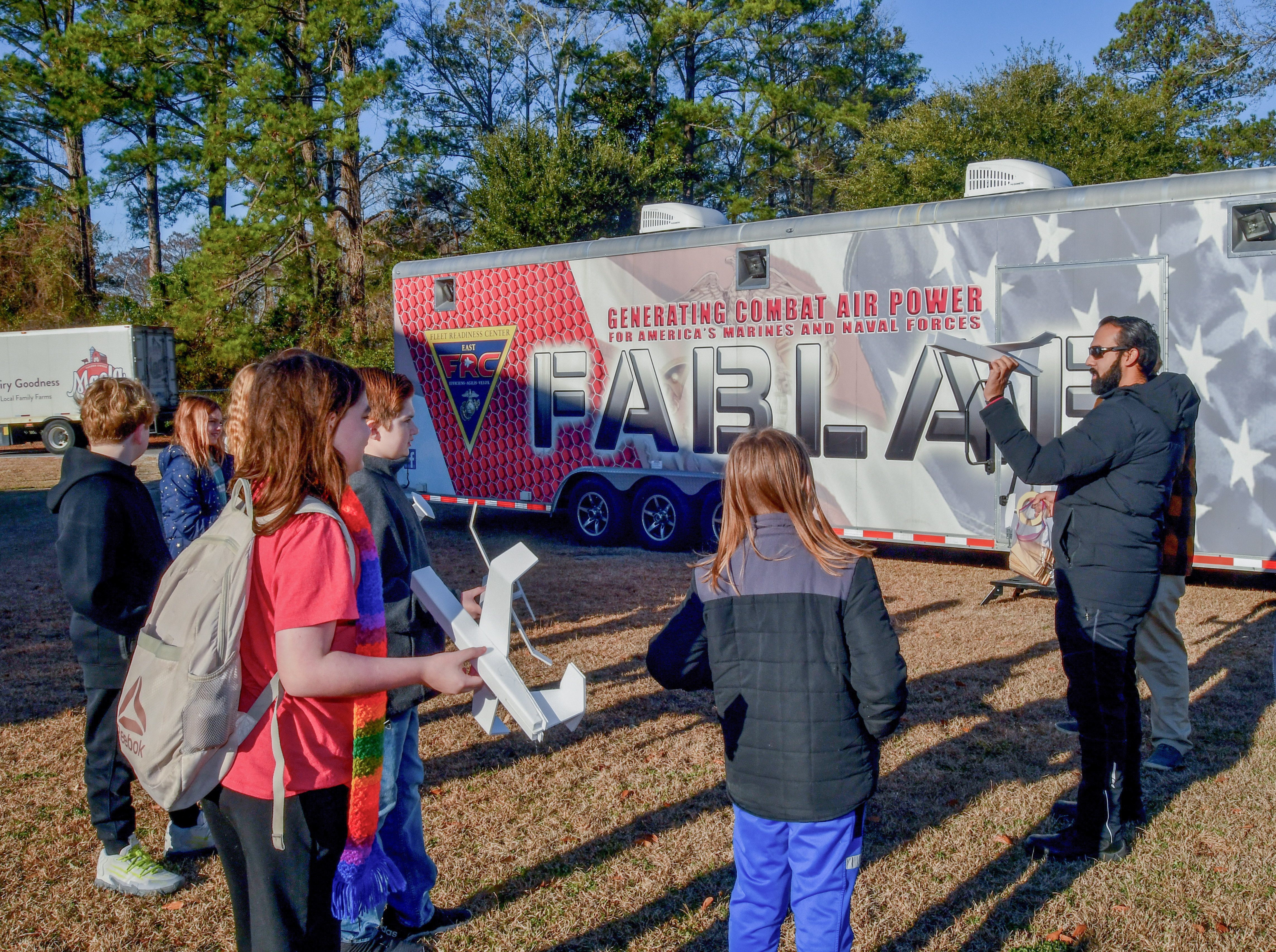 Man on the left stands in front of the FABLAB trailer holding a foam glider airplane and children stand on the left watching him