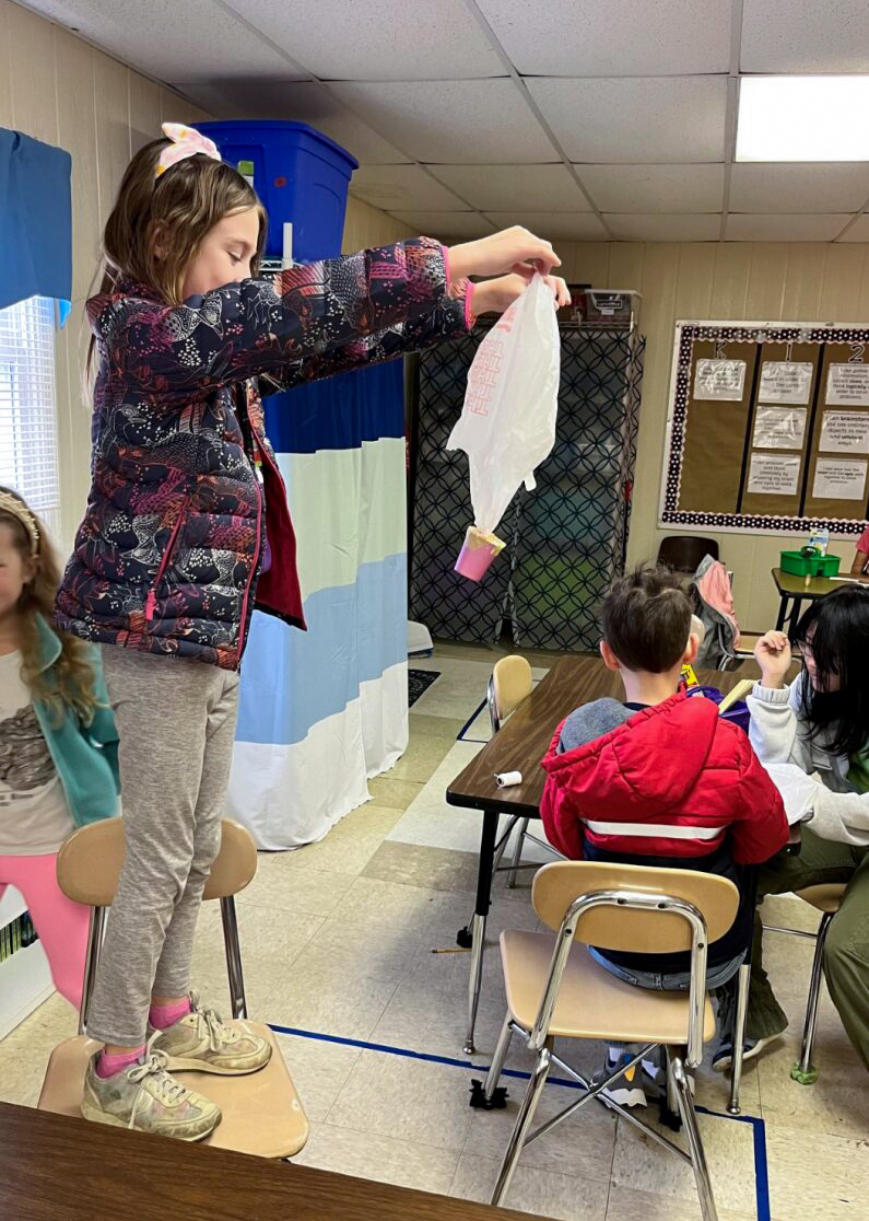 Student stands on top of chair holding a project made with a plastic bag 