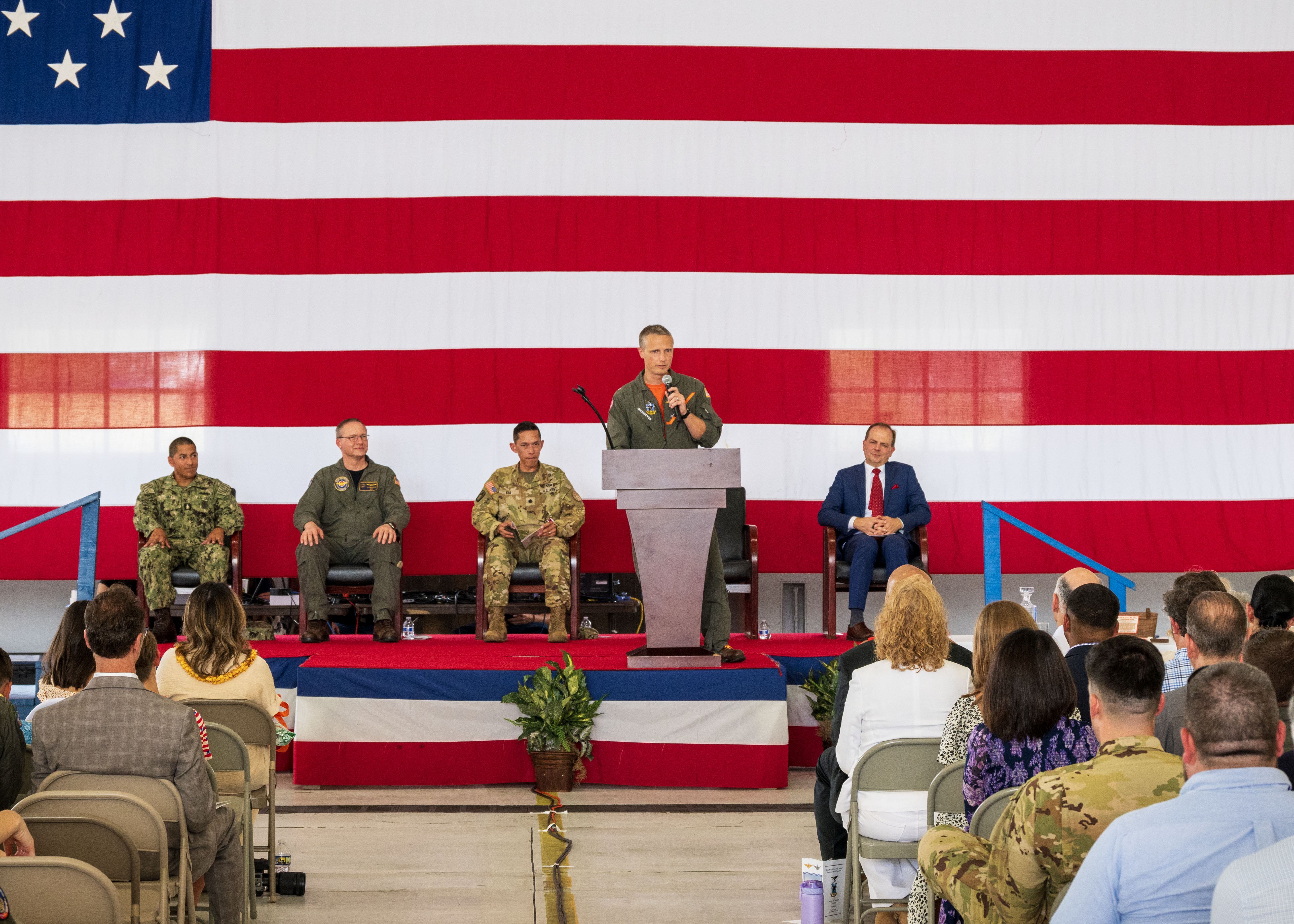 A military aviator stands behind a podium delivering remarks in on a stage in hangar spaces.