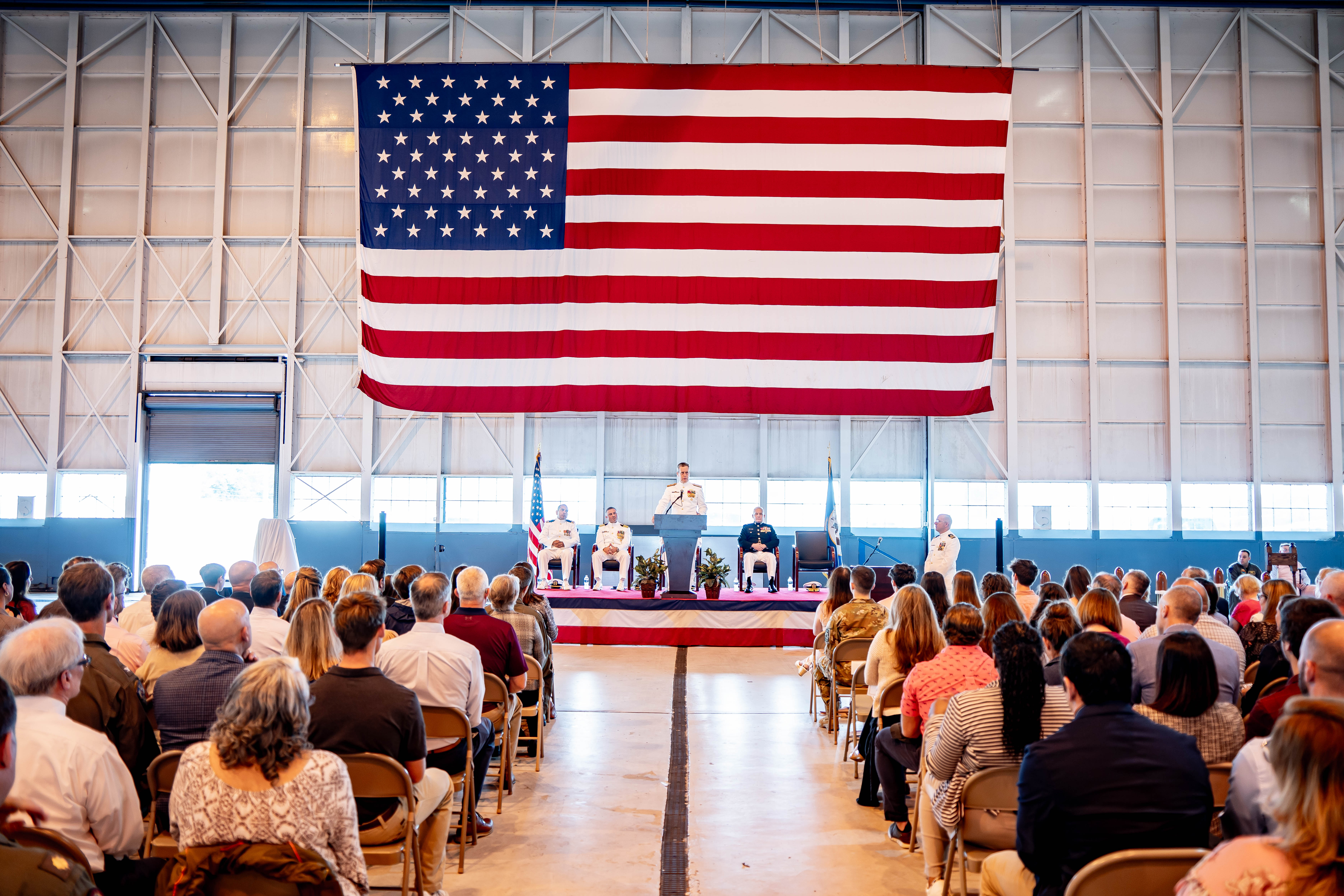A crowd sitting on chairs with a man on a stage speaking and a large USA flag behind