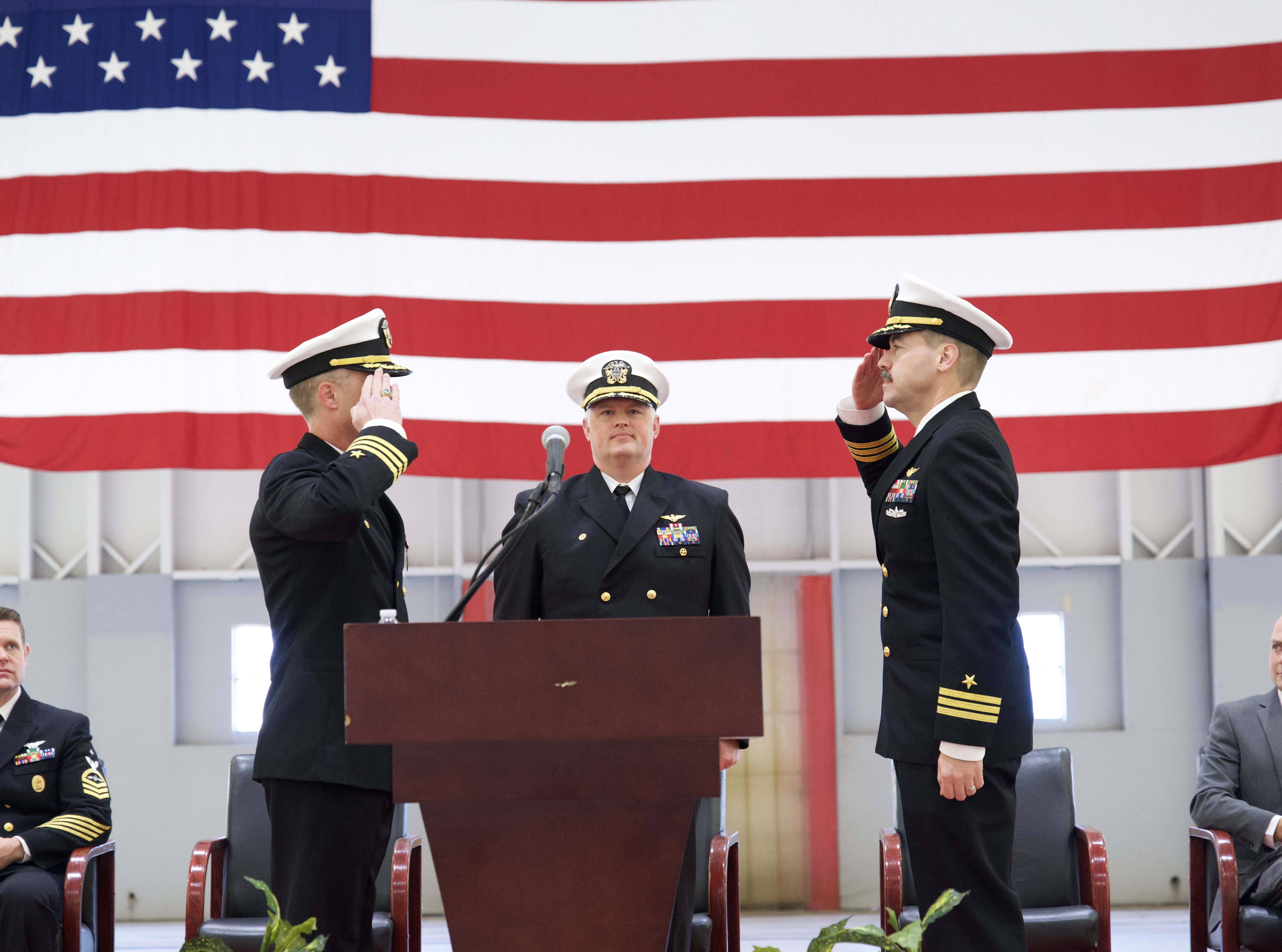 Naval officers salute each other in front of a podium during a ceremony inside a hangar.