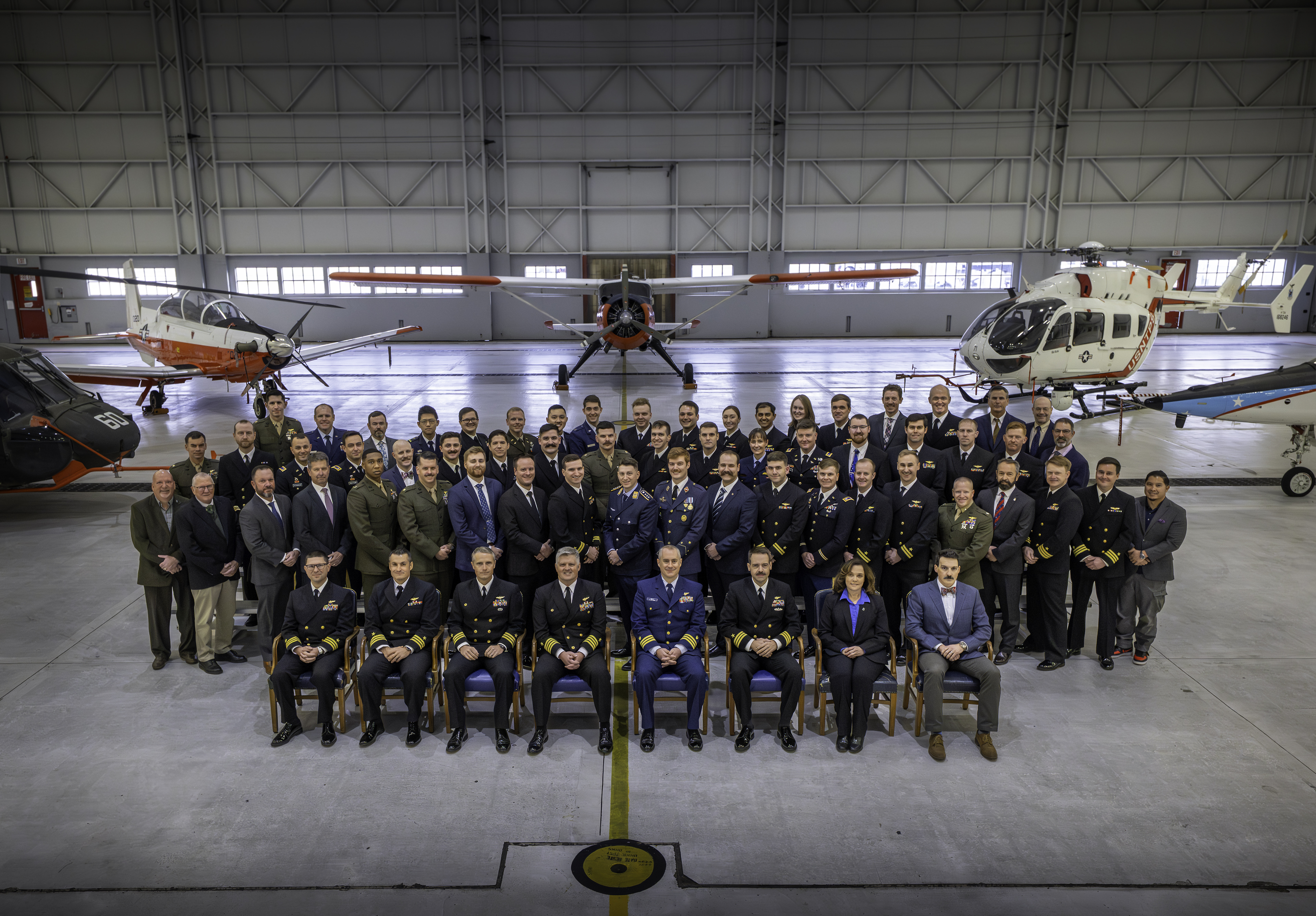 Four rows of people pose for a picture in front of five aircraft. The first row is seated, the back three rows are standing. Most are wearing their military dress uniform. There are a few civilians in business attire.