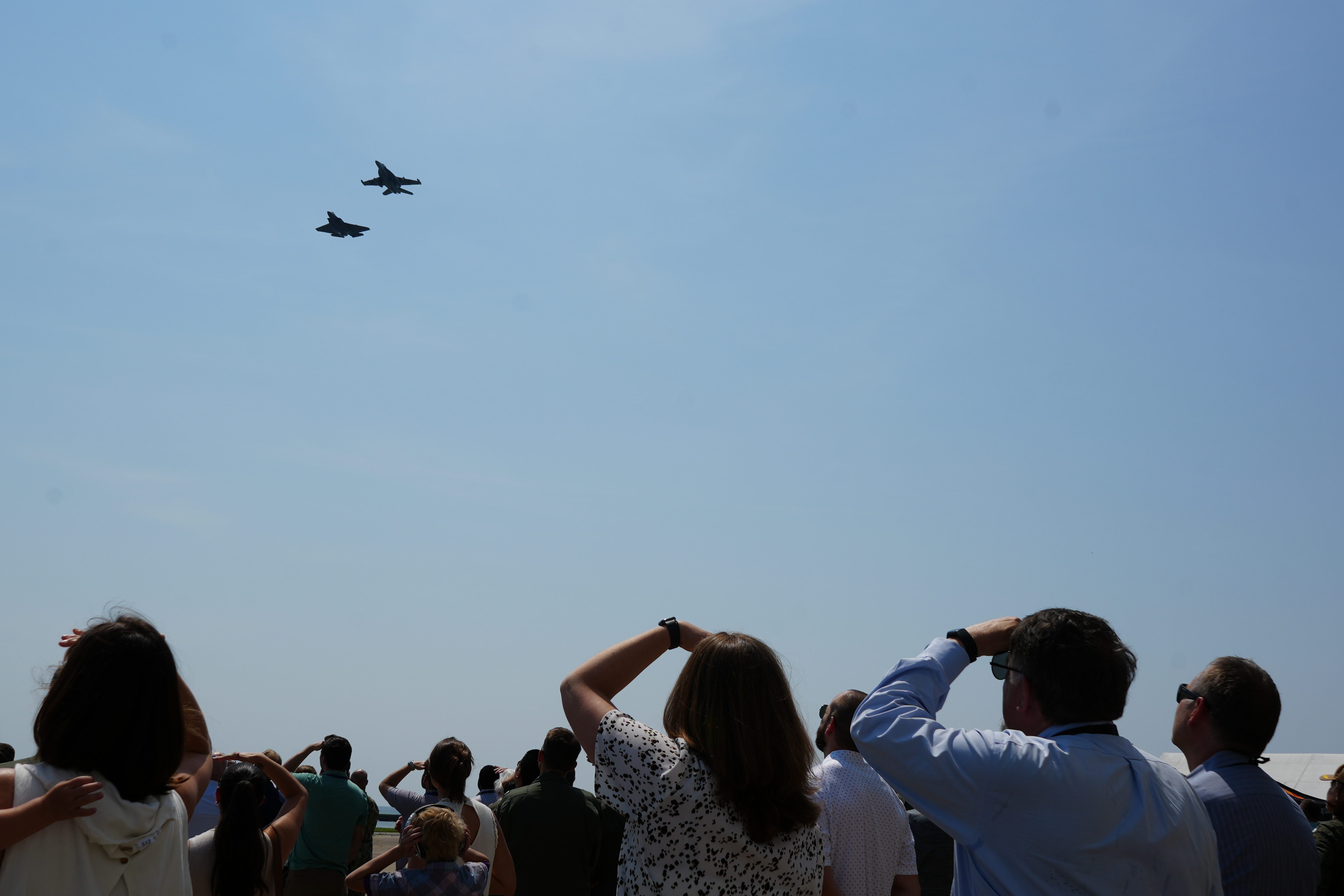 Two jets fly against clear blue skies while change of command ceremony attendees watch from the flightline. 