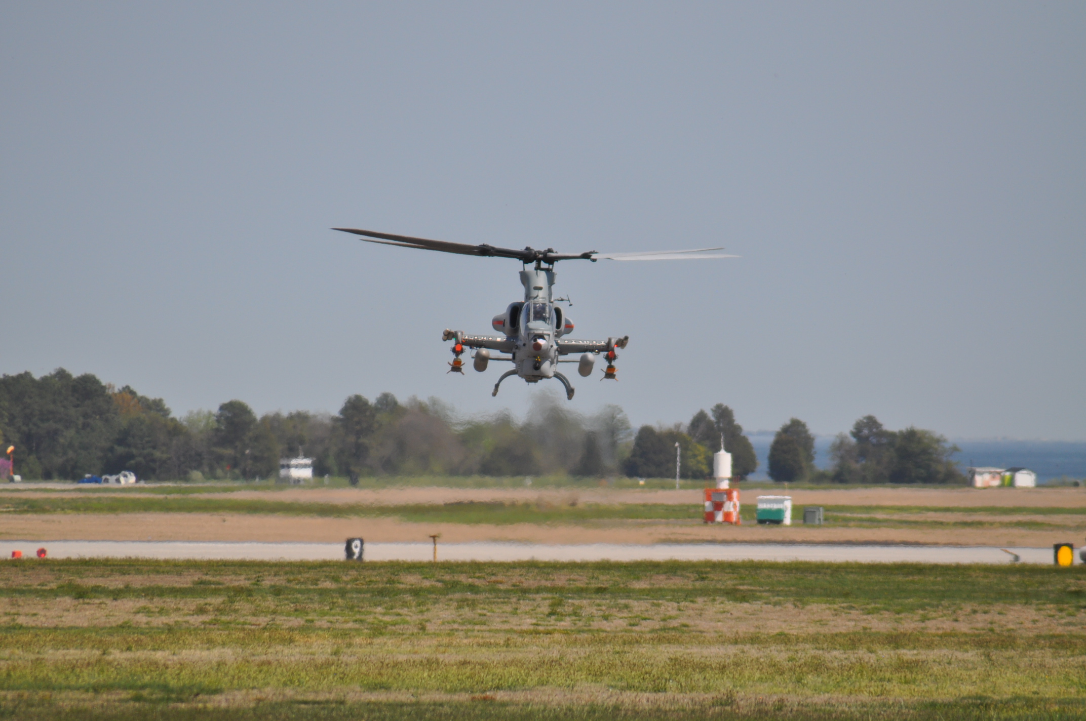 Helicopter hovering over a green lawn and part of a runway with trees in the background