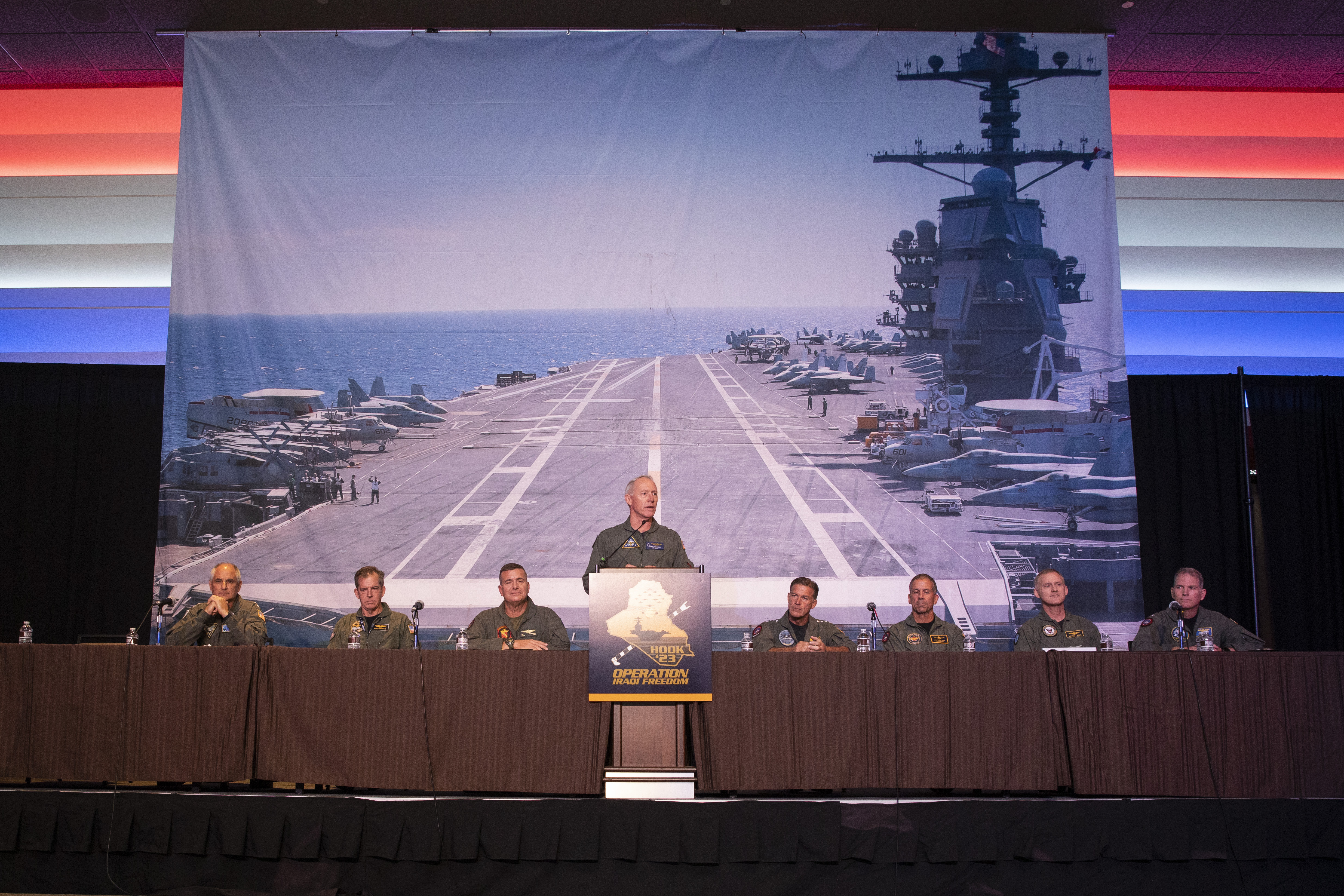 Man in flight suit stands at podium, men in flight suits sit at long table, photo of aircraft carrier deck behind them.