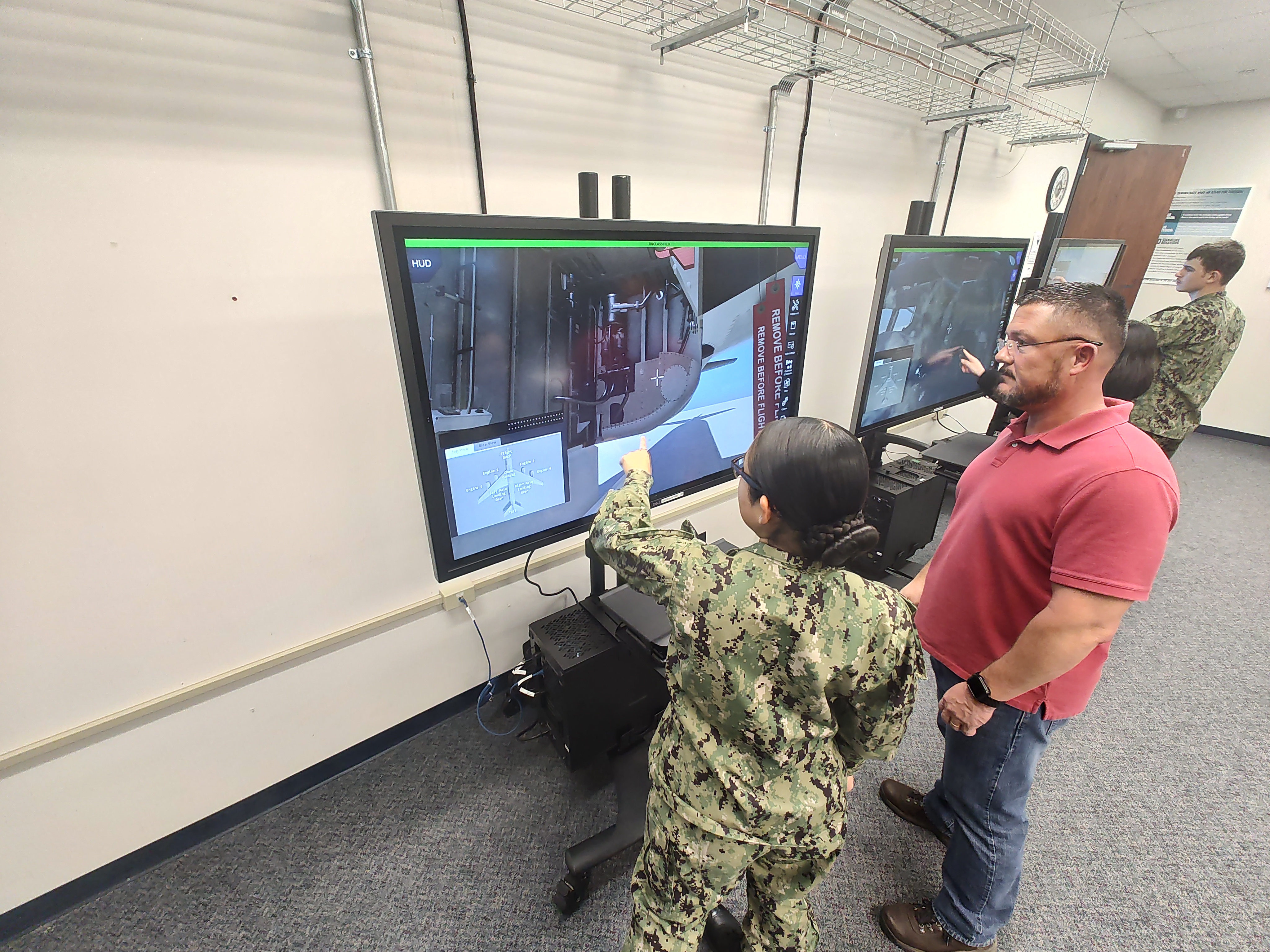 Sailor in uniform conducts training on a virtual touch-screen while being supervised by an instructor in a red shirt