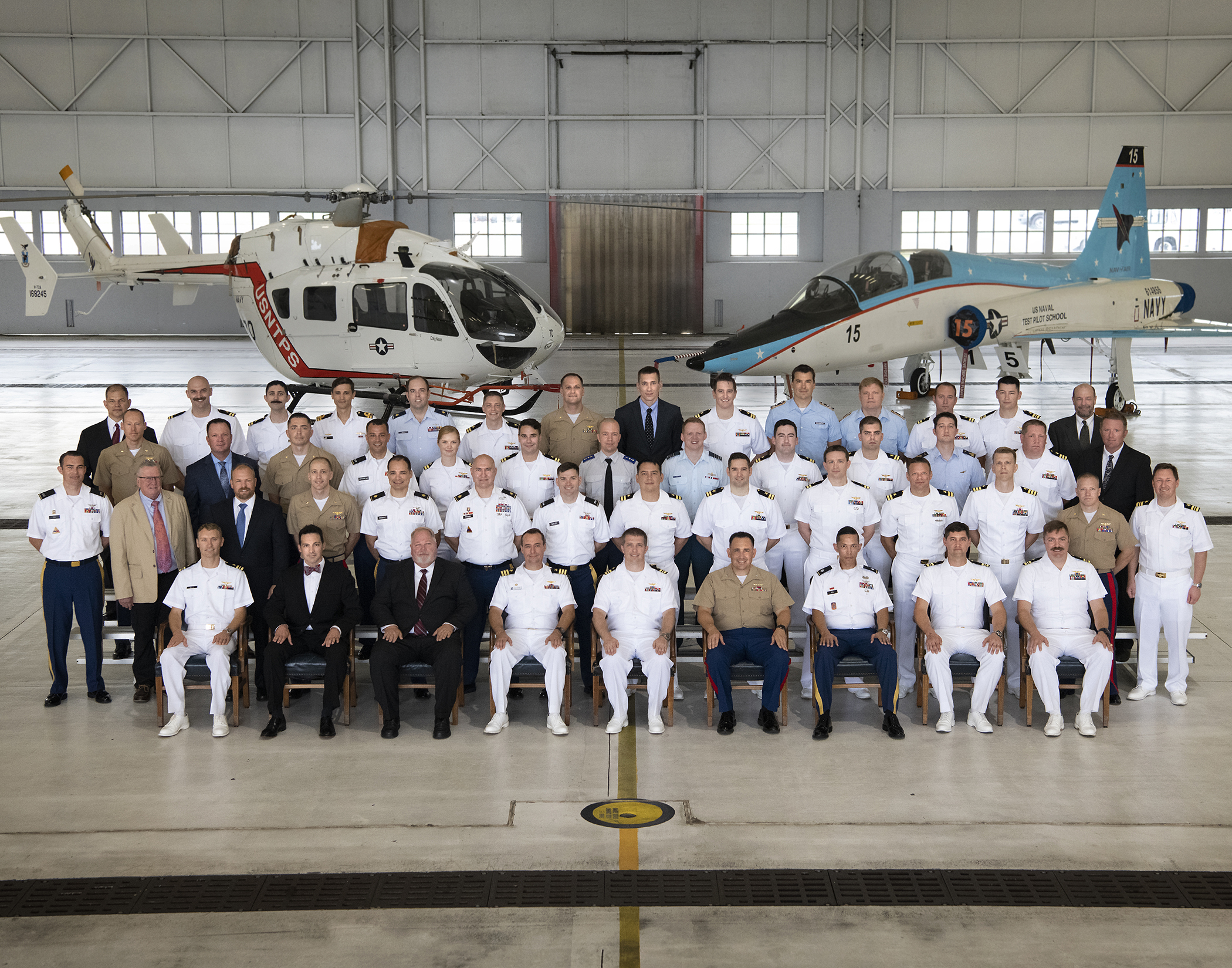 Military and civilian test pilots in uniform and formal attire pose for a class photo inside a hangar with a helicopter and jet. 