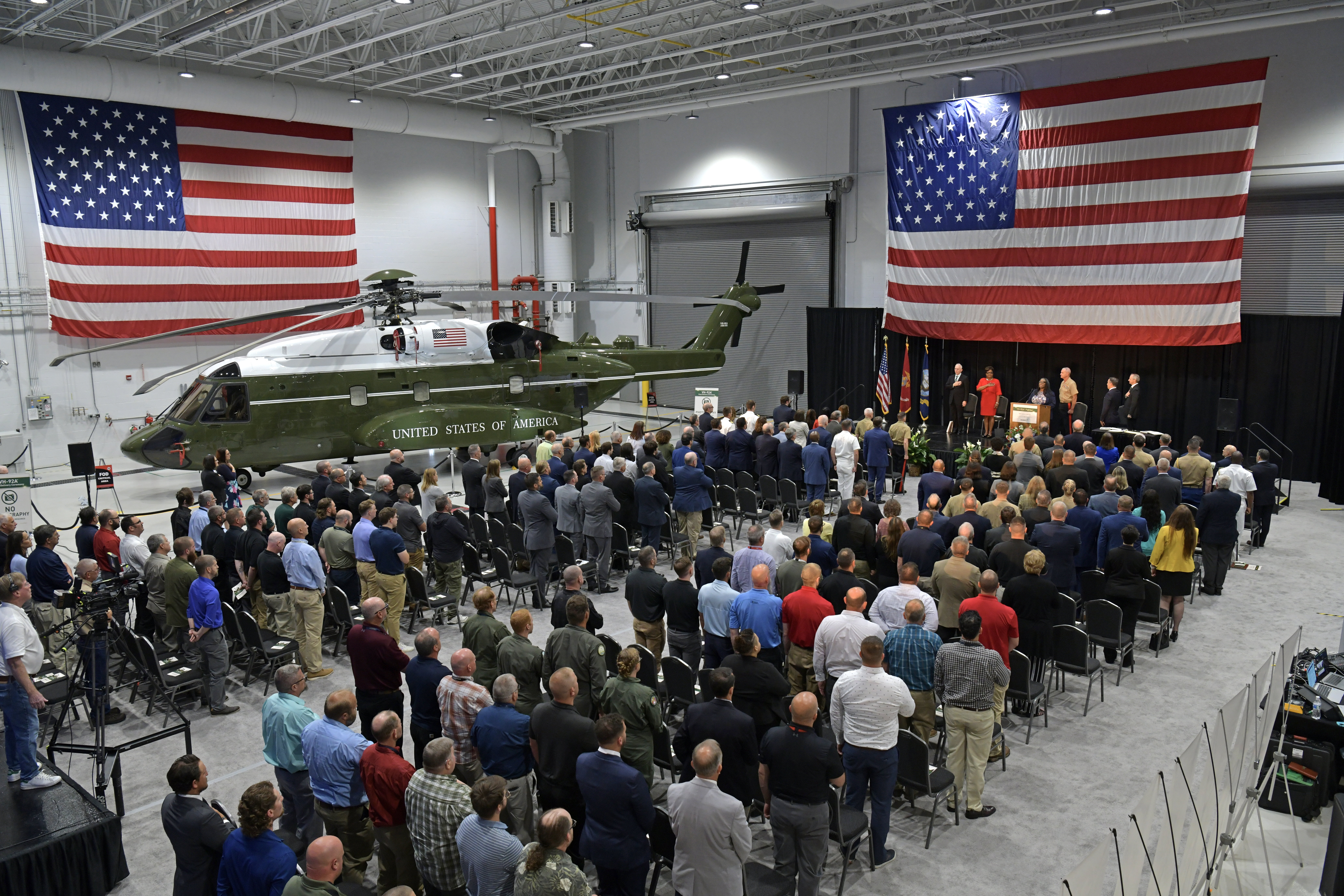 People seated in rows with a stage in front and a helicopter and two American flags 