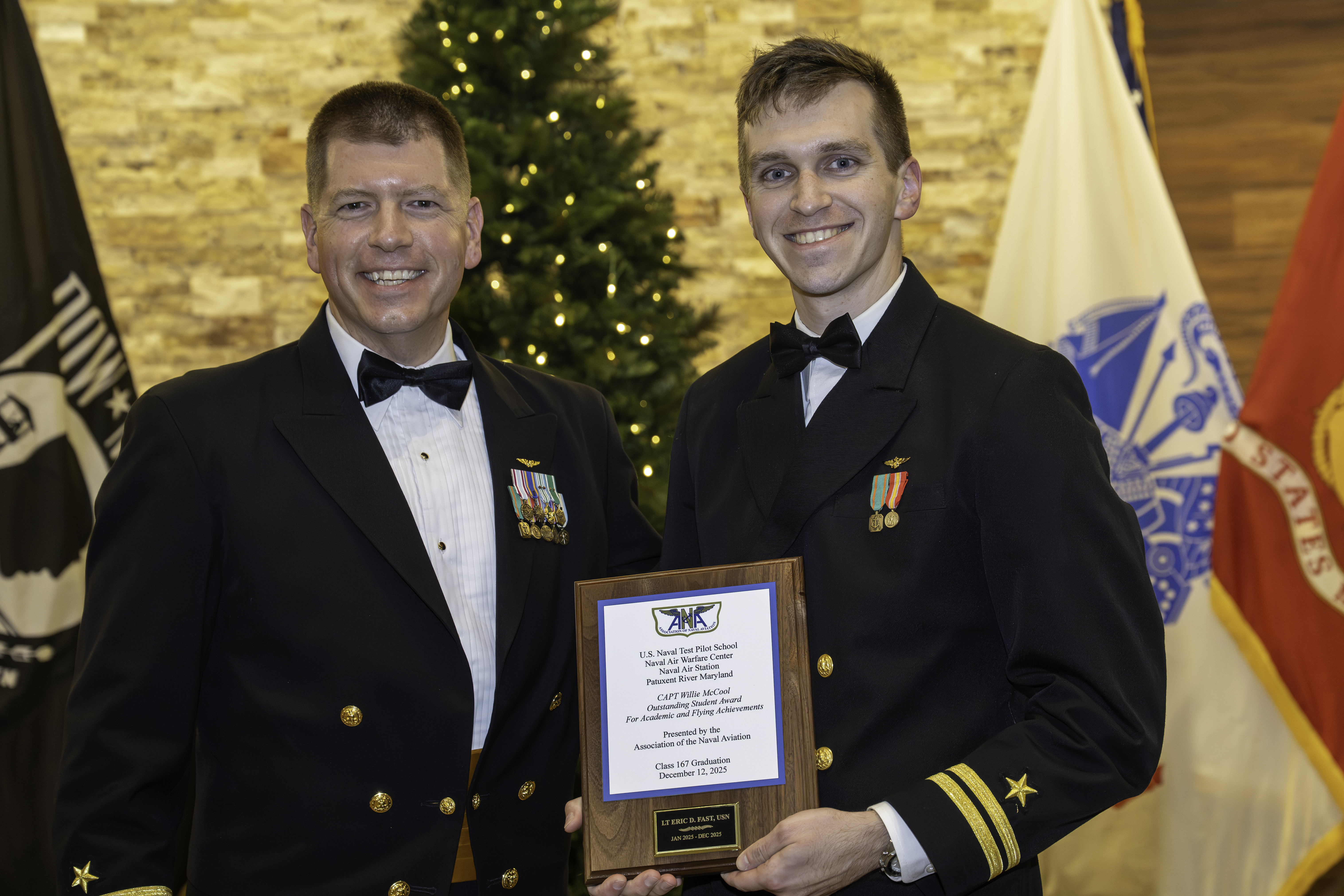 Two Naval Officers pose in front of flags. The one on the left is holding a plaque.