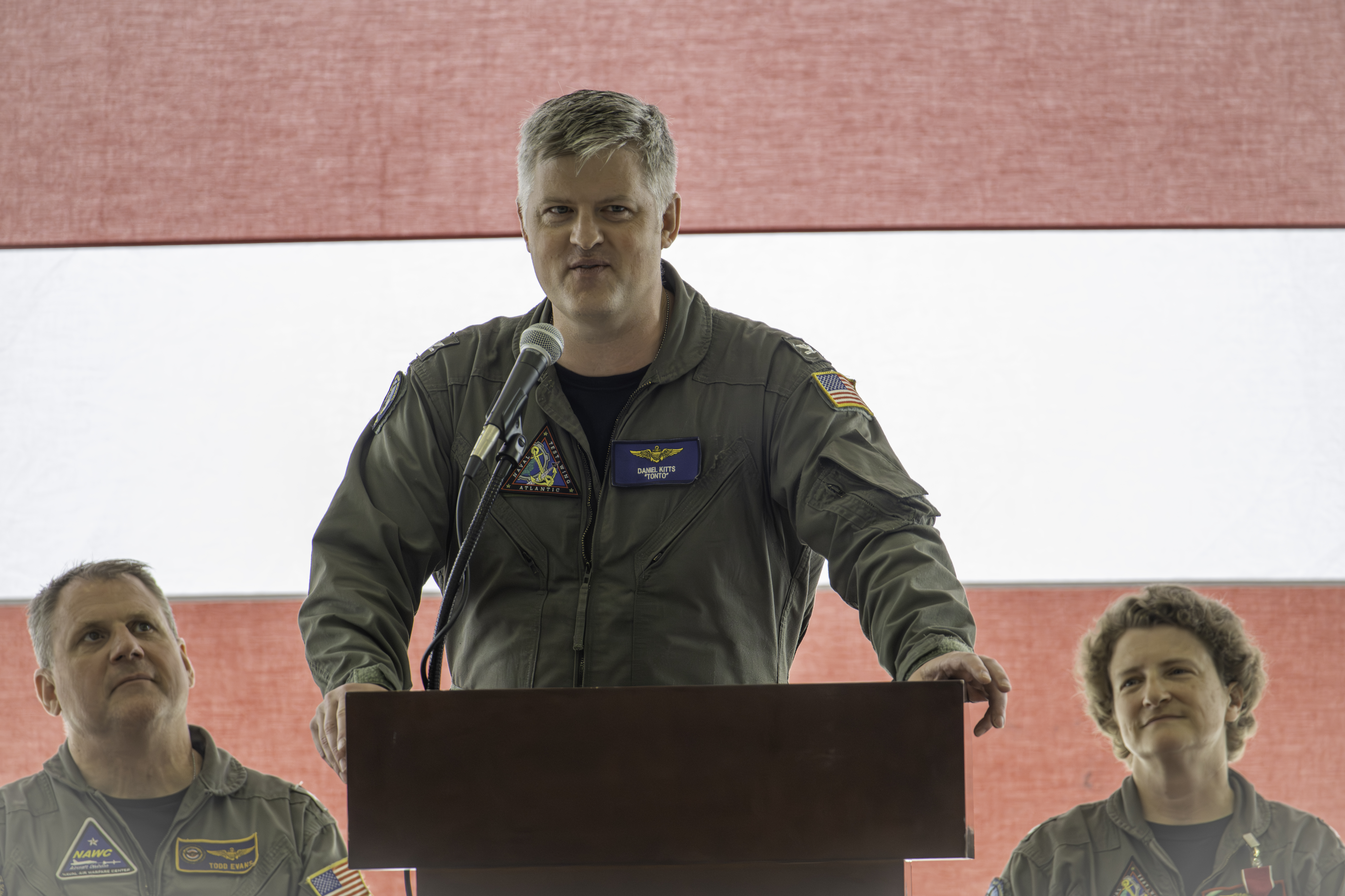 The Naval Test Wing Atlantic Commodore addresses ceremony attendees in a hangar at a podium.