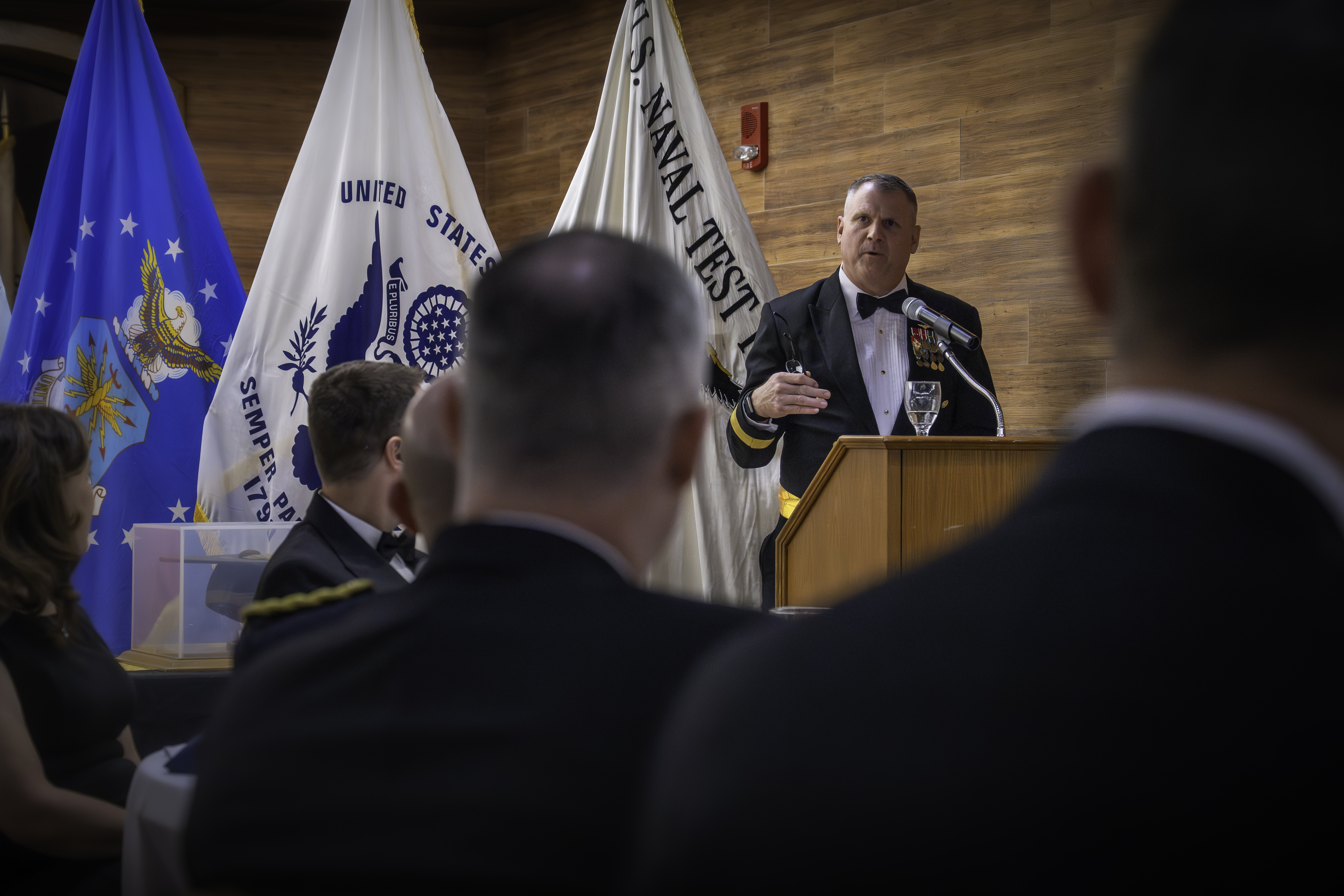With the backs of attendee's heads in the foreground, a Navy Rear Admiral Speaks at a wooden podium in front of three flags. 