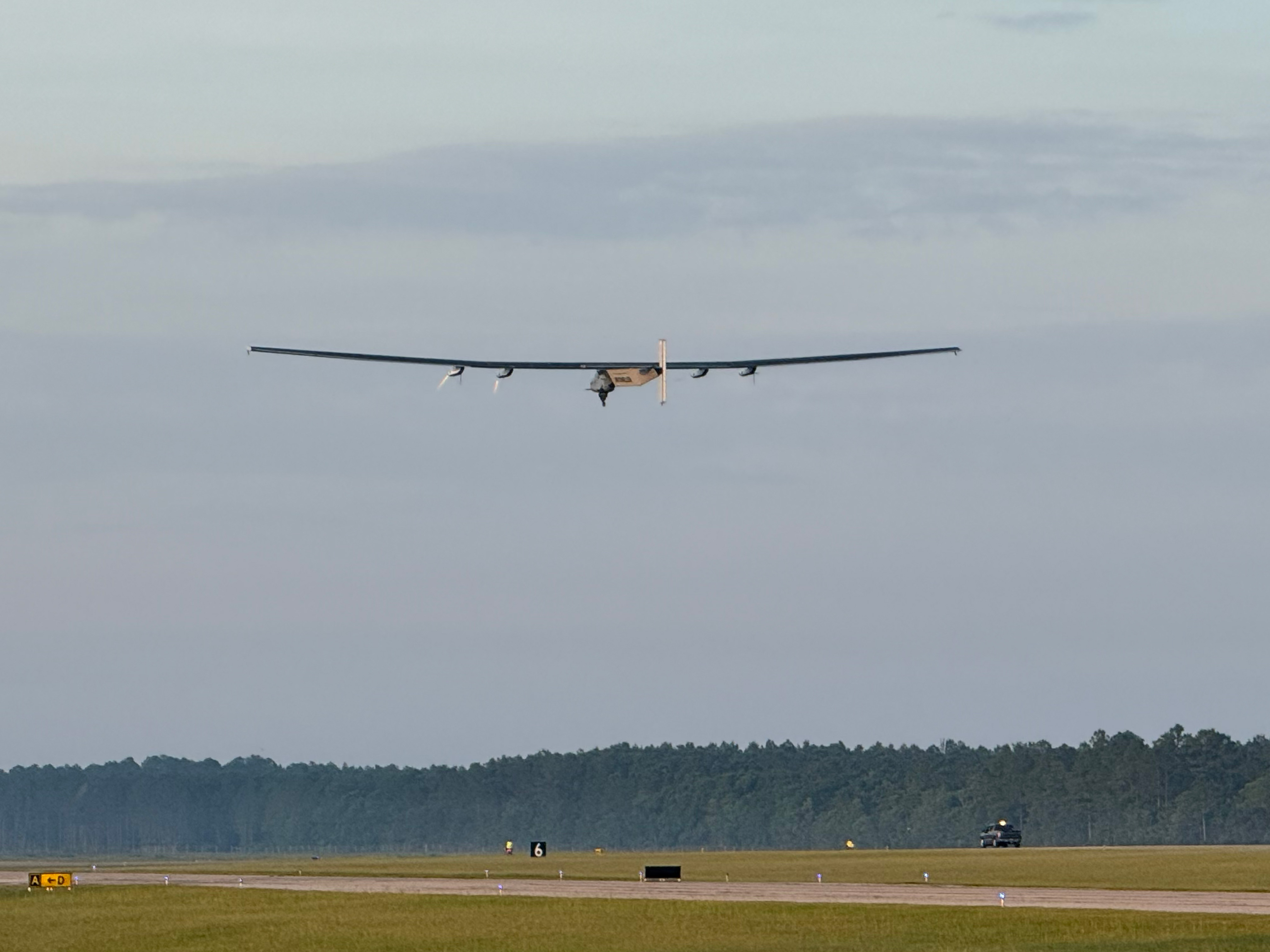 A large unmanned aerial vehicle is flying low to the ground above a runway. There are trees in the background.