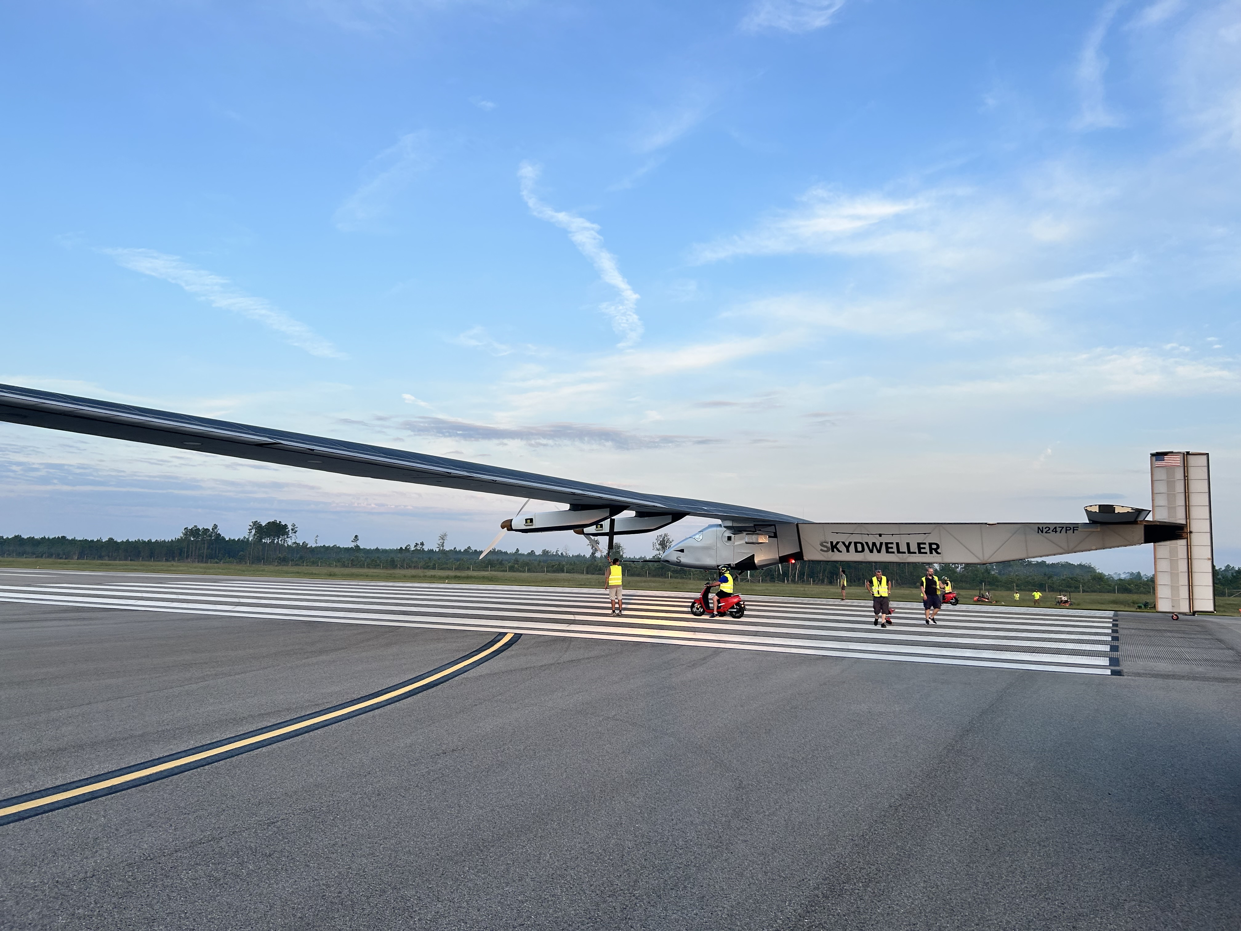 A large unmanned aerial system is pictured on a runway prior to takeoff. There are four people in the picture who are preparing the UAS for takeoff.