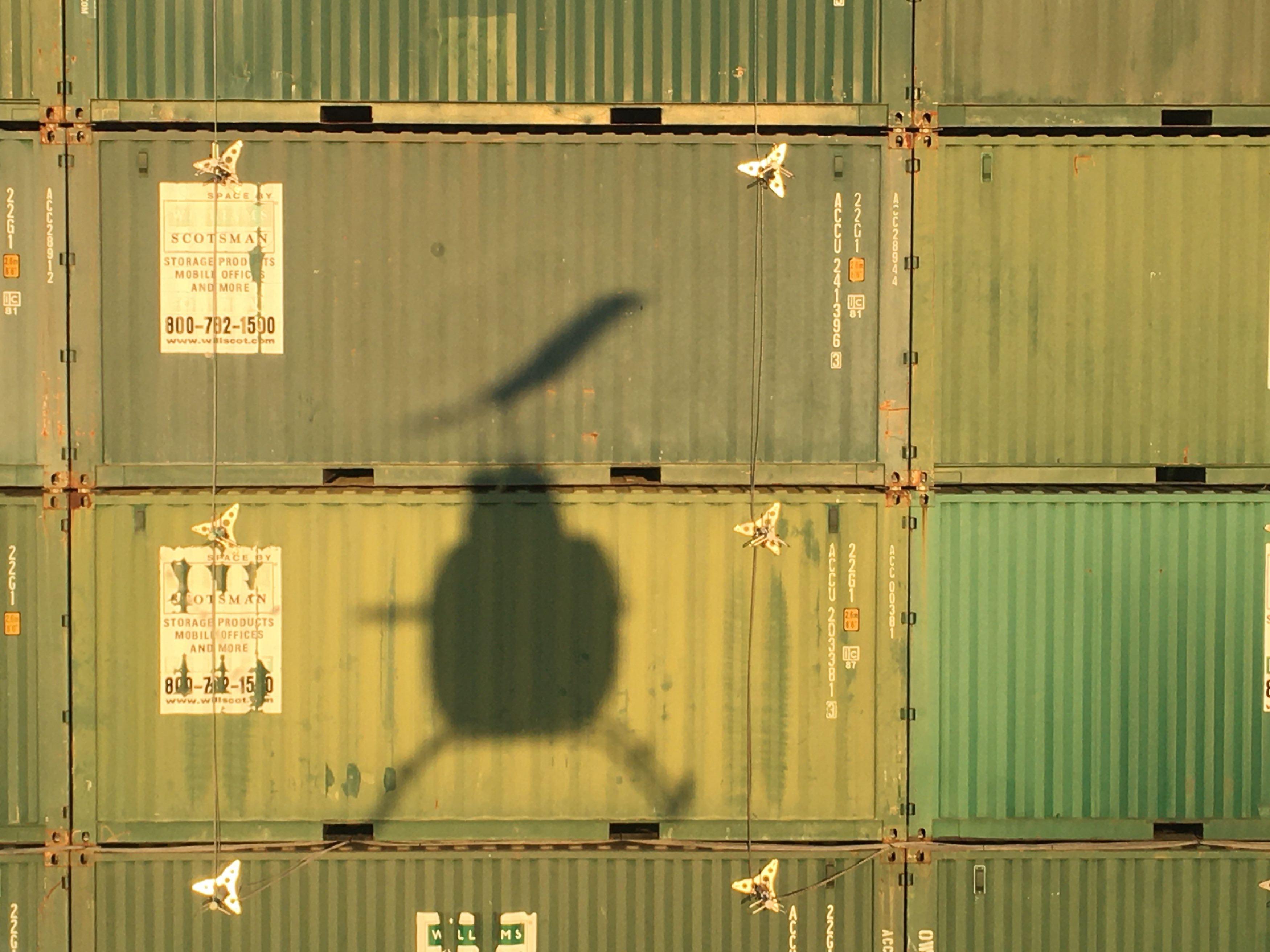 The shadow of a U.S. Naval Test Pilot School (USNTPS) OH-58C Kiowa falls on the face of the Elevated Fixed Platform, a test platform constructed from over 100 shipping containers stacked to form a 38-foot-tall box and capped with steel plates, which USNTPS test pilots recently used to evaluate the hover performance of helicopters near obstacles and inside confined areas.