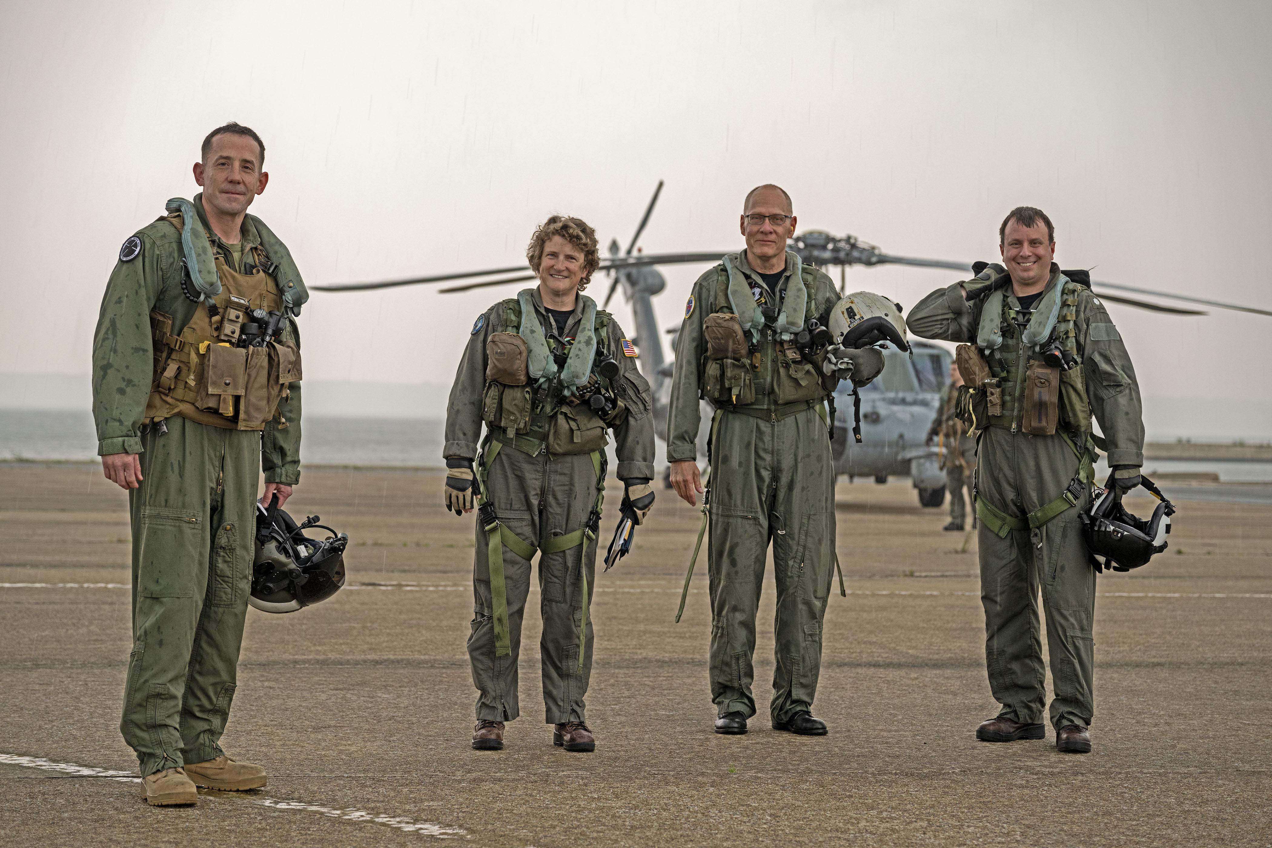 Aviators in flight suits stand in front of a helicopter on a flight line against a smoky horizon on the Patuxent River.