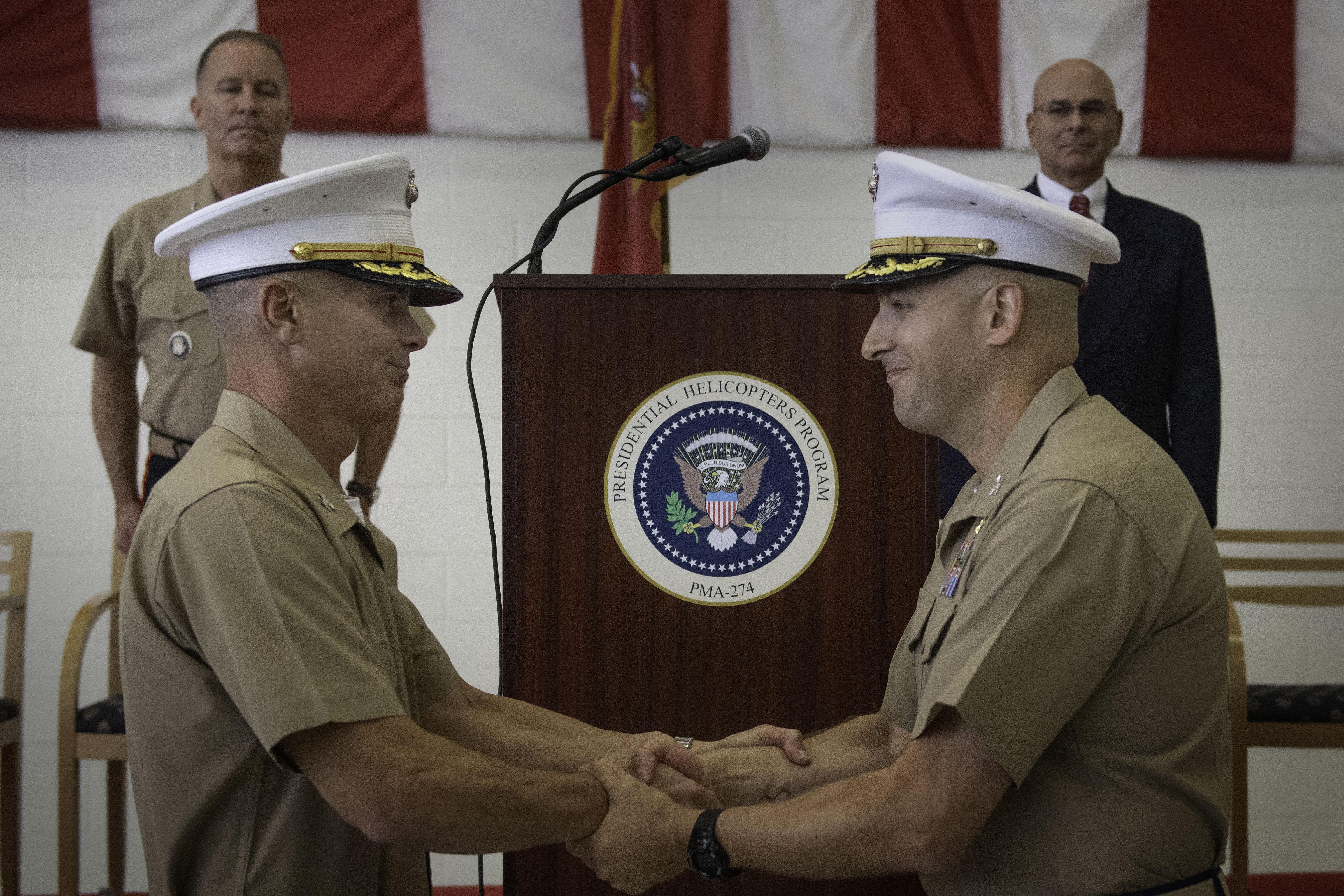Two marines shake hands after a change of command ceremony in front of a podium with the PMA-274 seal