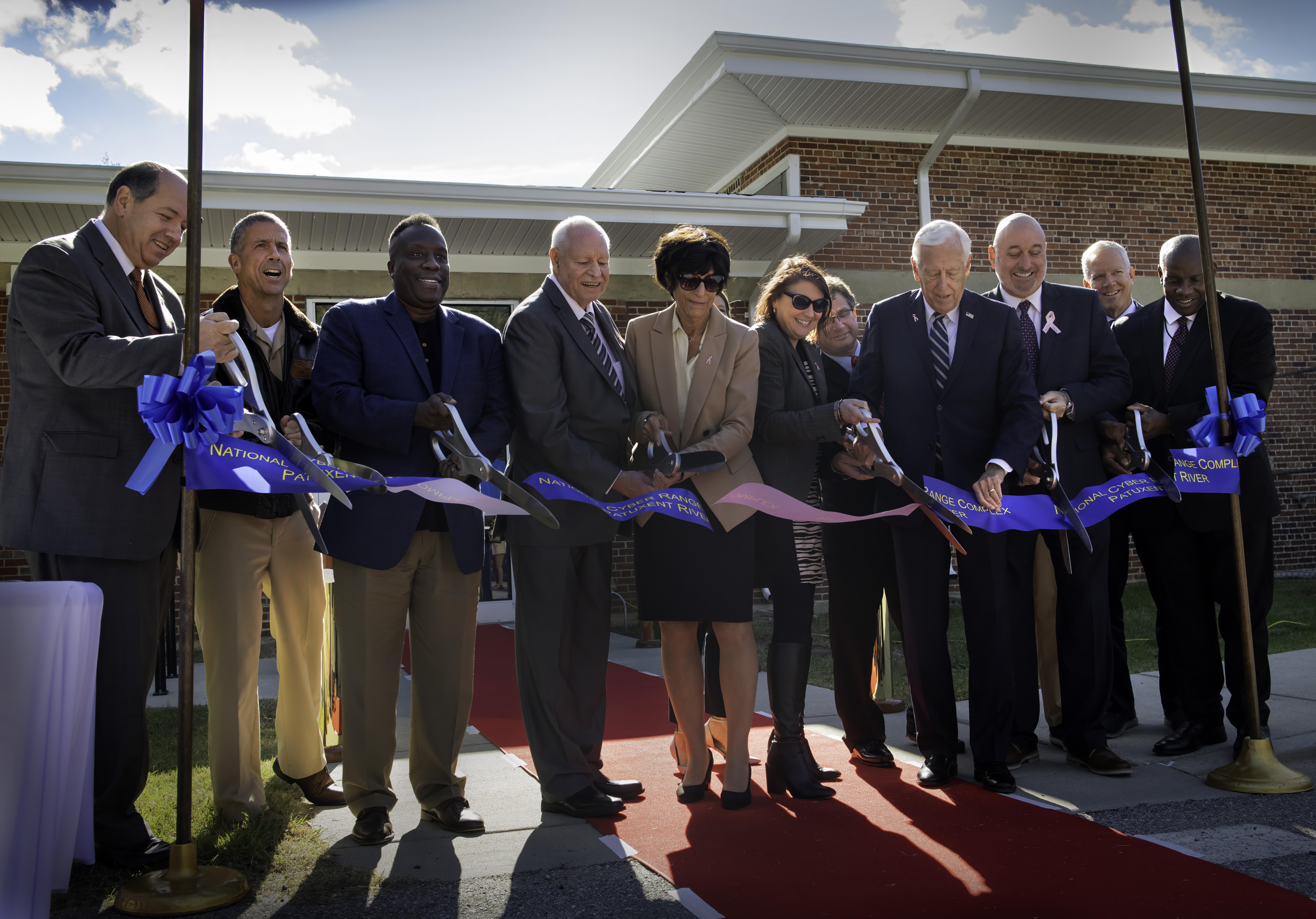 Professionals stand shoulder-to-shoulder and cut a blue ribbon founding a new test facility under sunny skies.