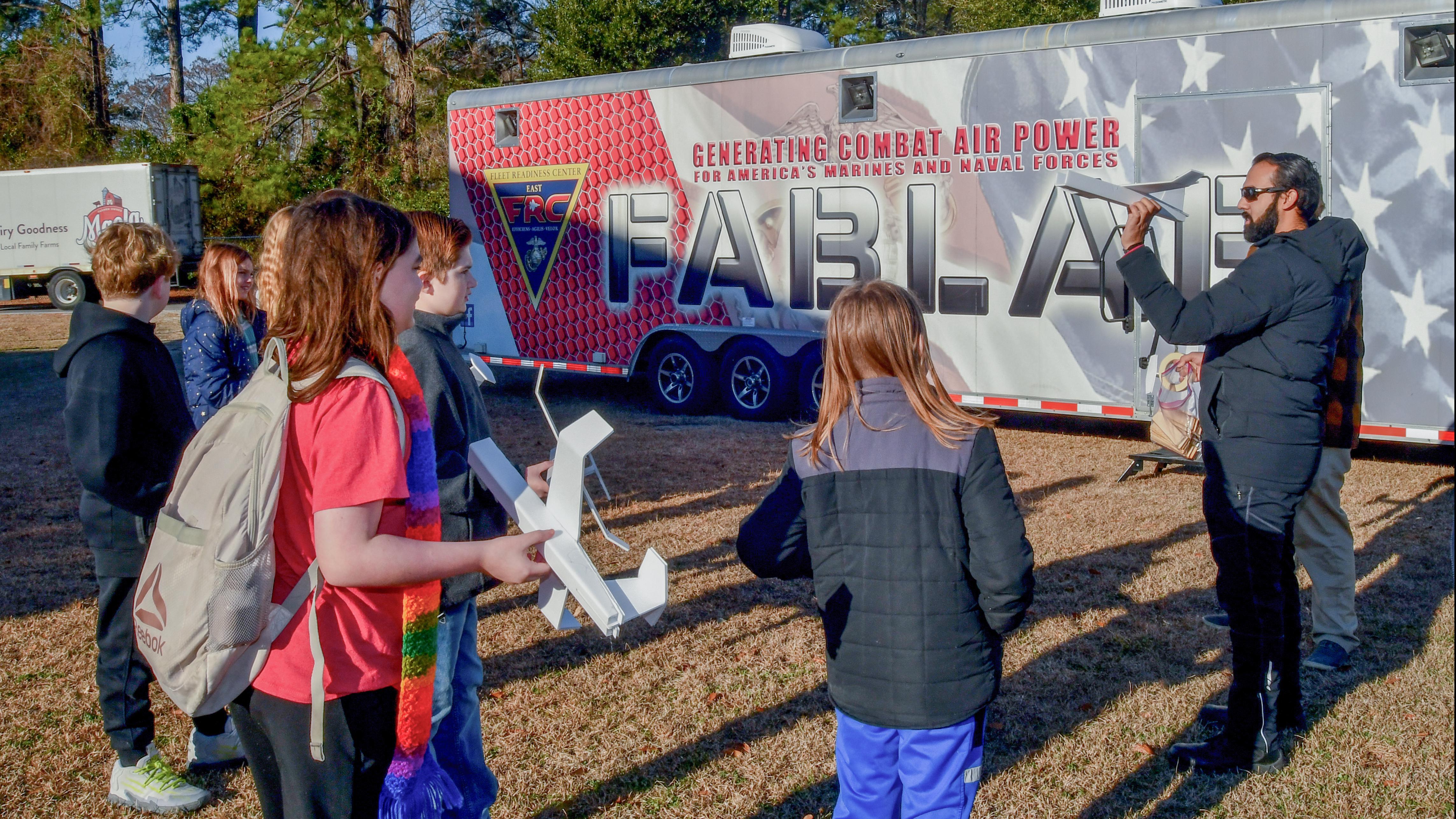 Man on the left stands in front of the FABLAB trailer holding a foam glider airplane and children stand on the left watching him