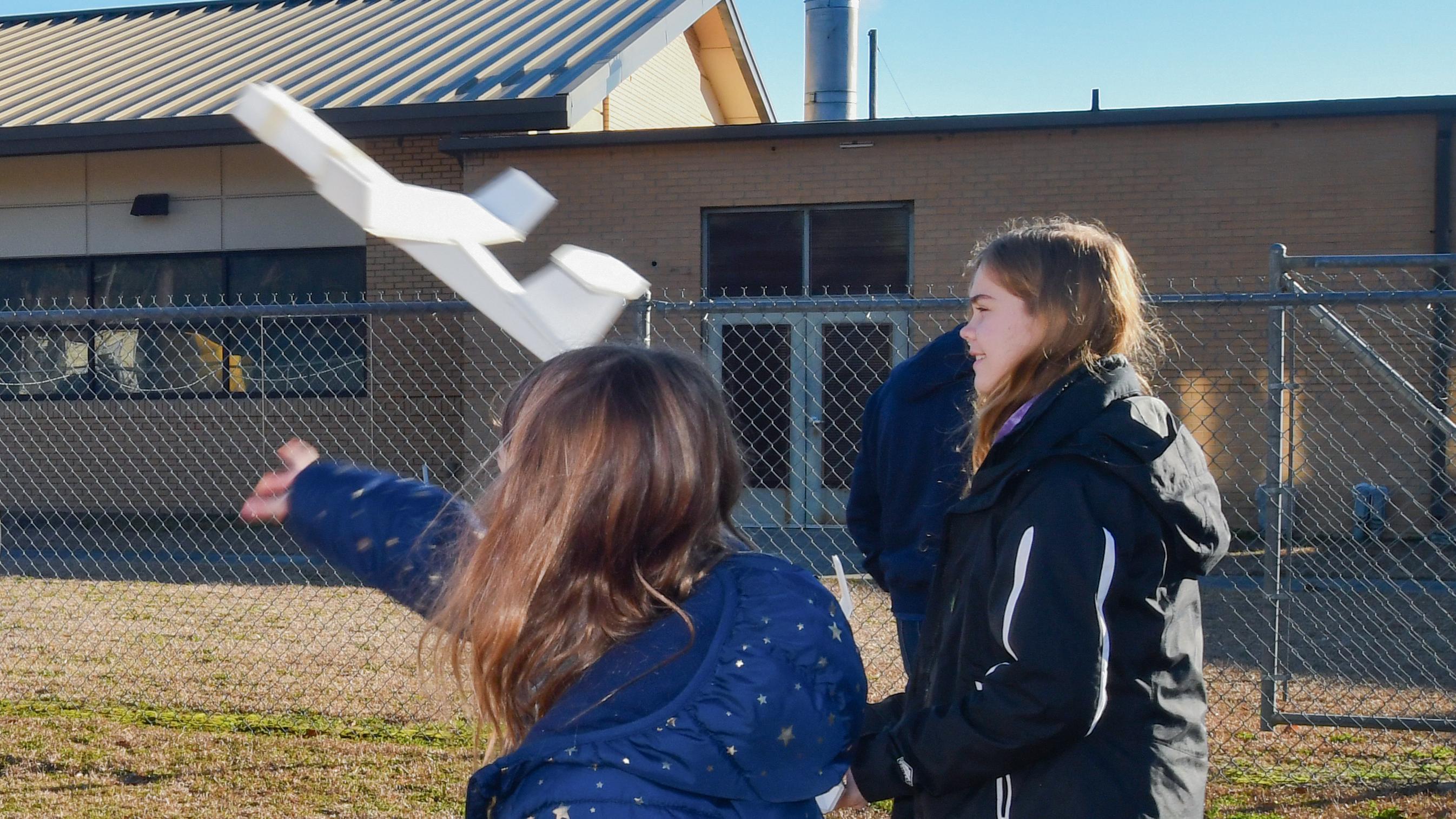 Two female students stand outside and one is throwing a foam airplane into the air
