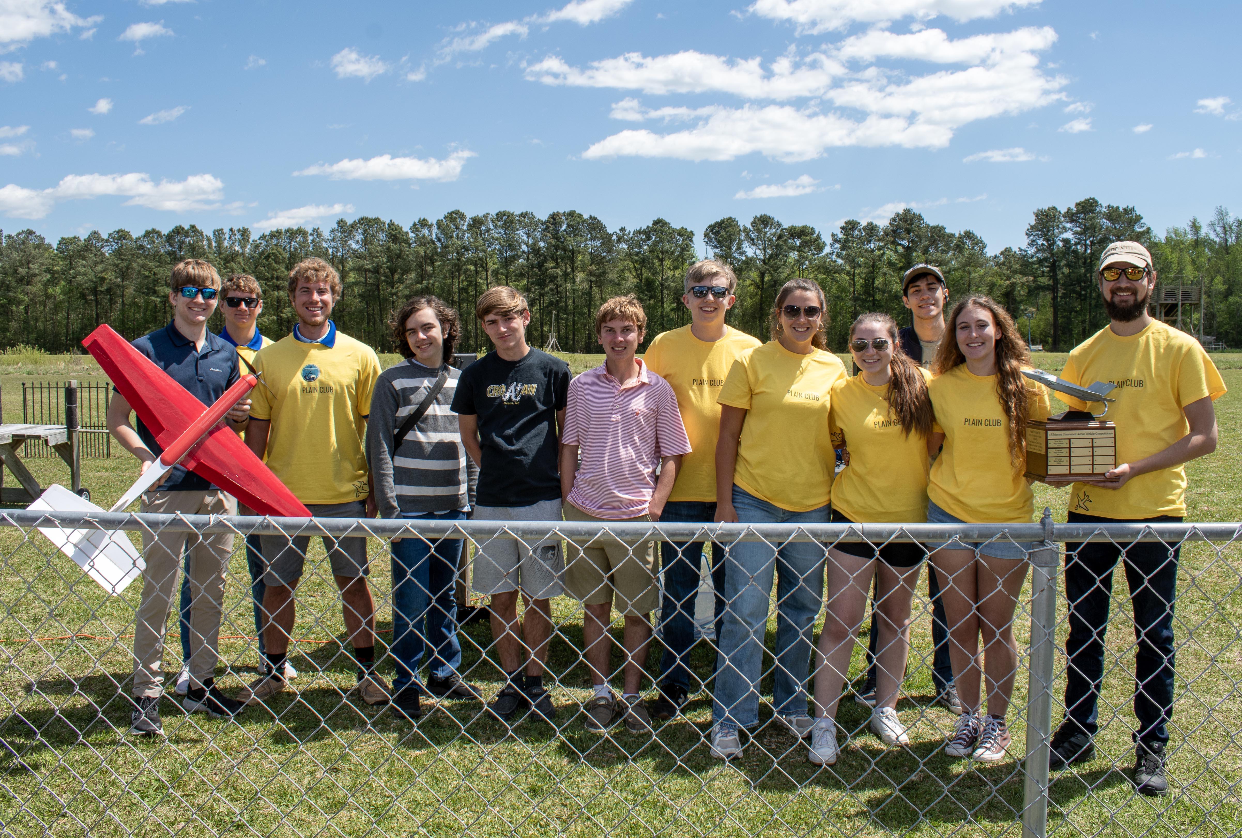 Team of students wearing yellow stand outside with trophy and model aircraft