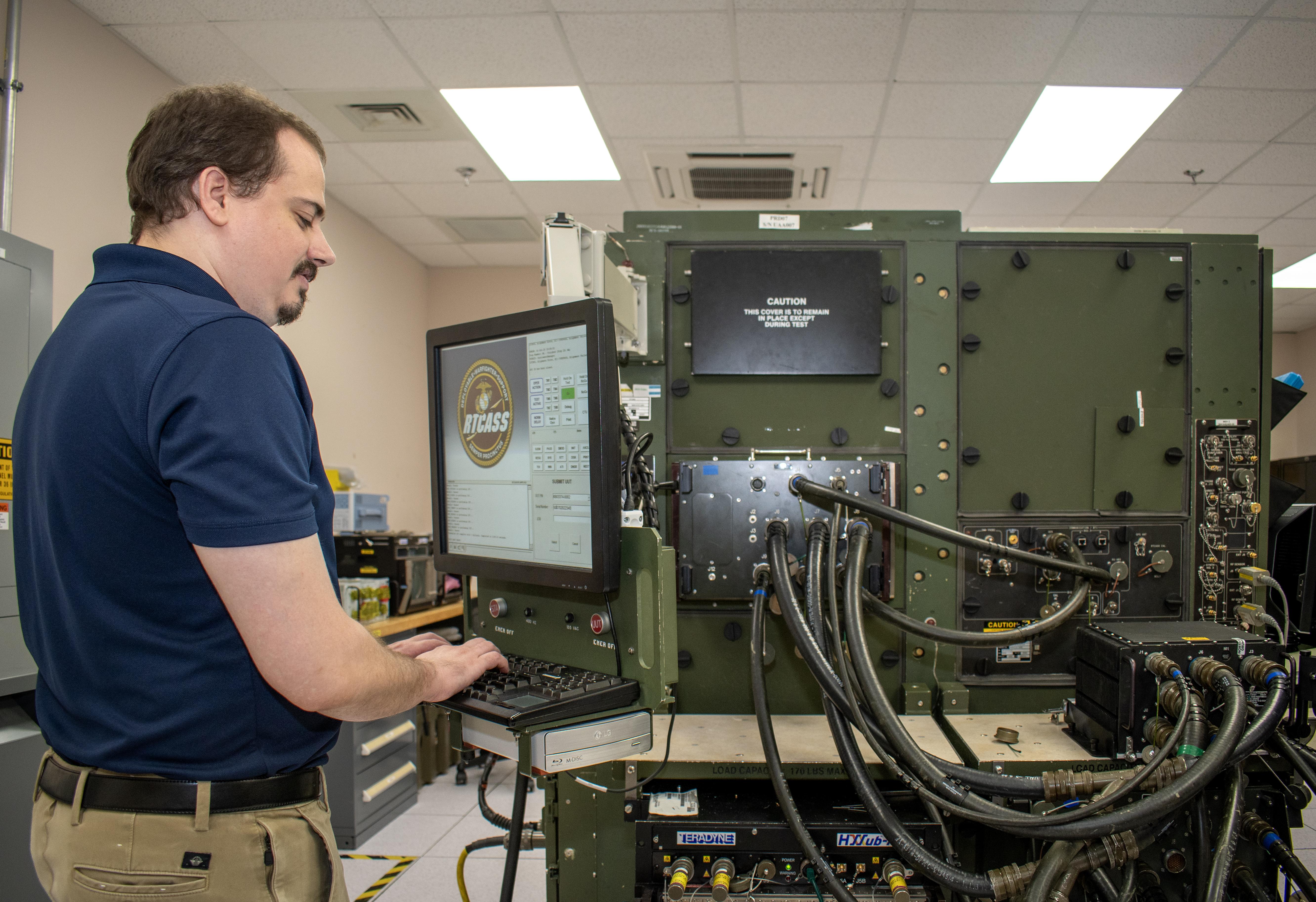 Man in blue shirt works at computer in front of large, green machine 