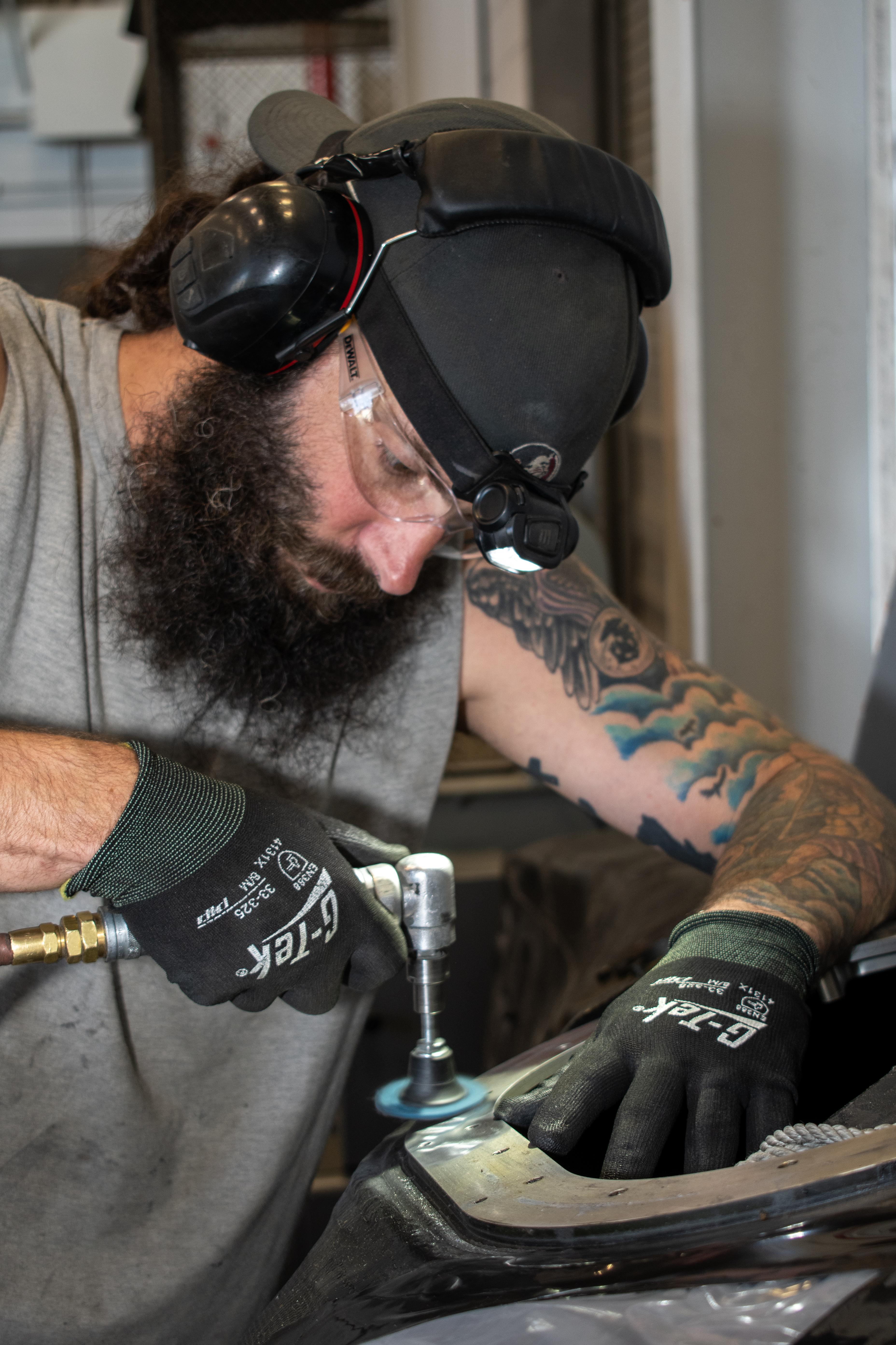 Male aircraft worker wearing personal protective safety equipment uses a hand tool to smooth a metal surface.