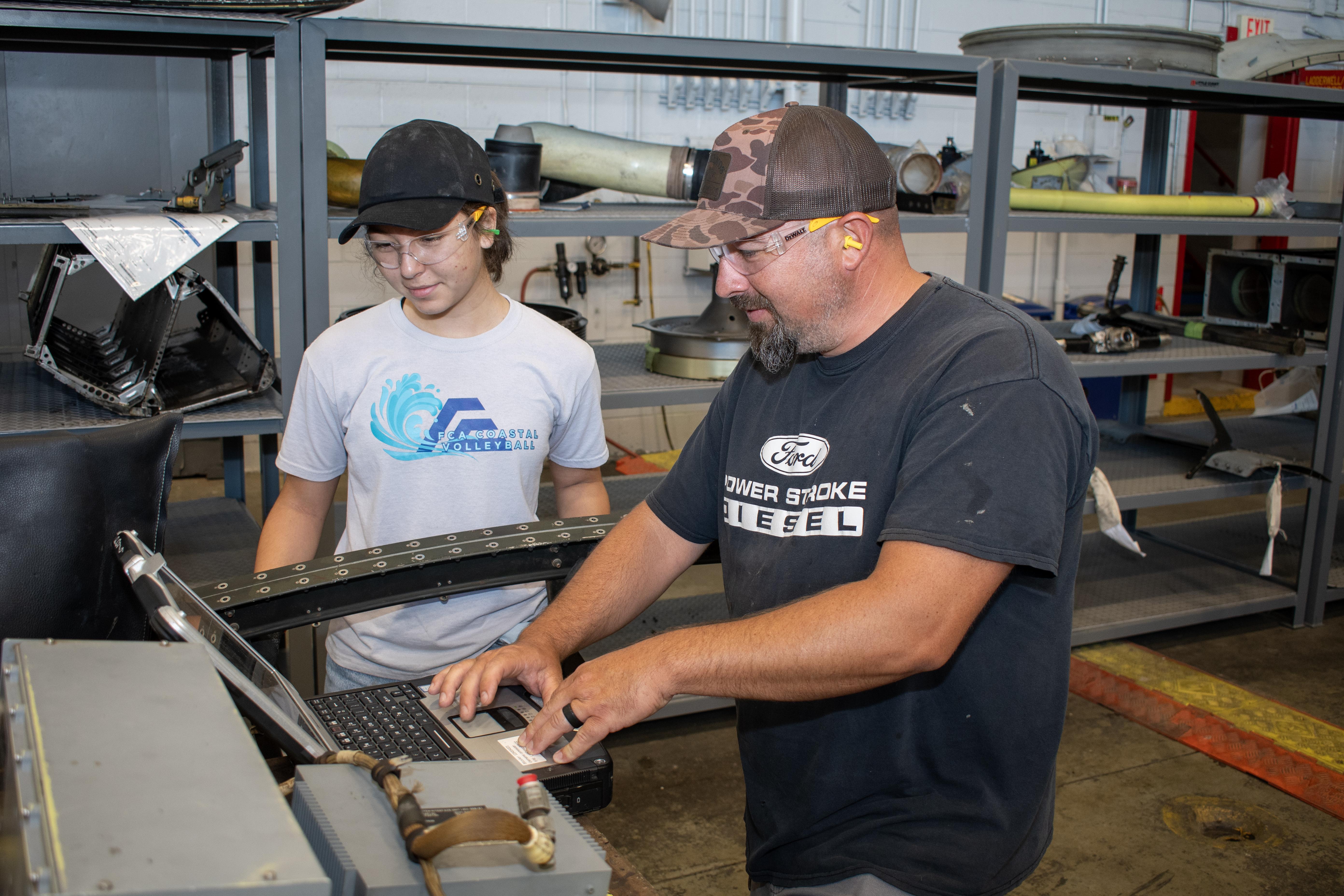 Male aircraft work leader demonstrates use of computer-based maintenance manual for high-school aged, female summer intern. 