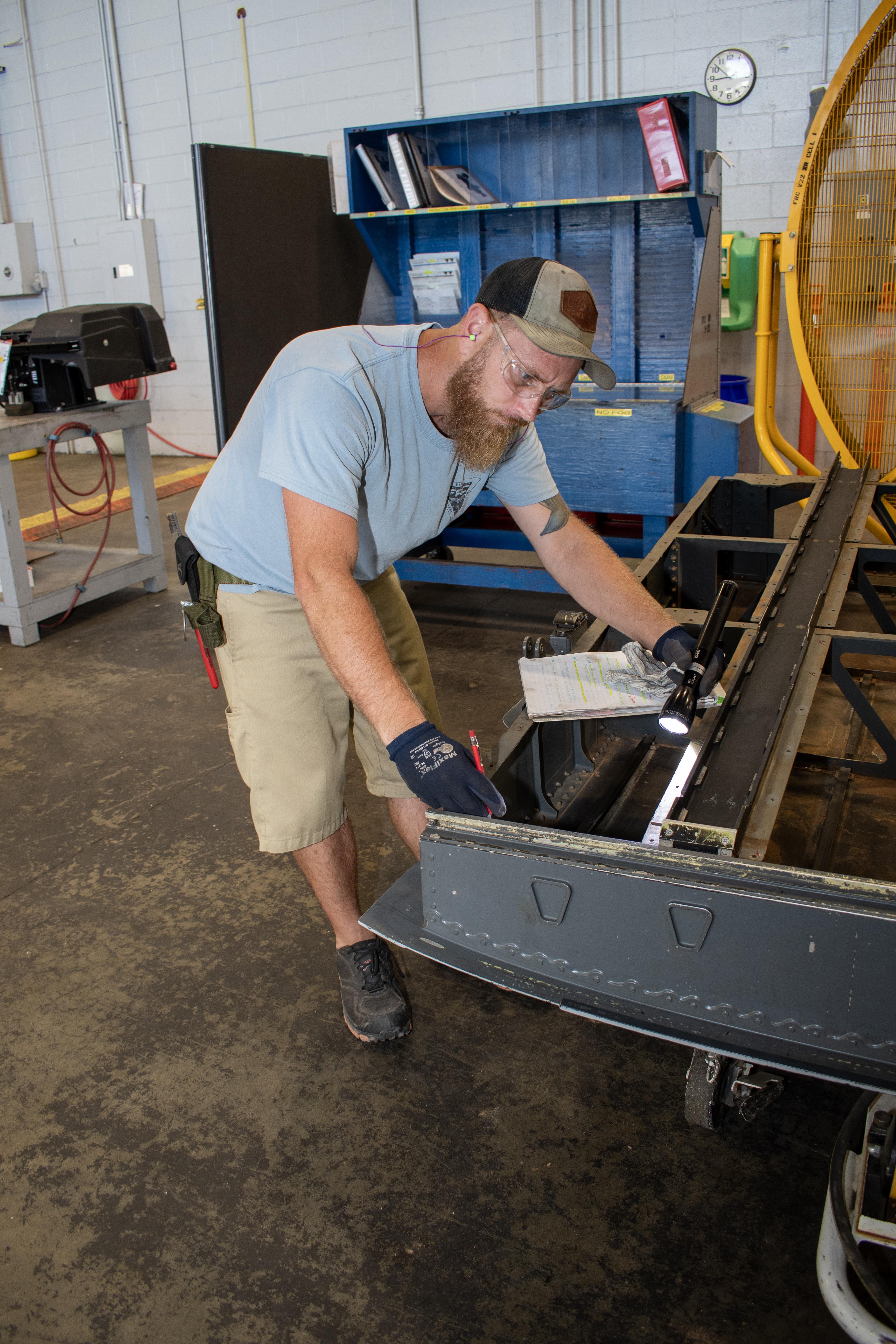 Bearded male aircraft evaluator wearing personal protective safety equipment uses a flashlight to inspect the ramp of a V-22 Osprey for potential maintenance requirements. 