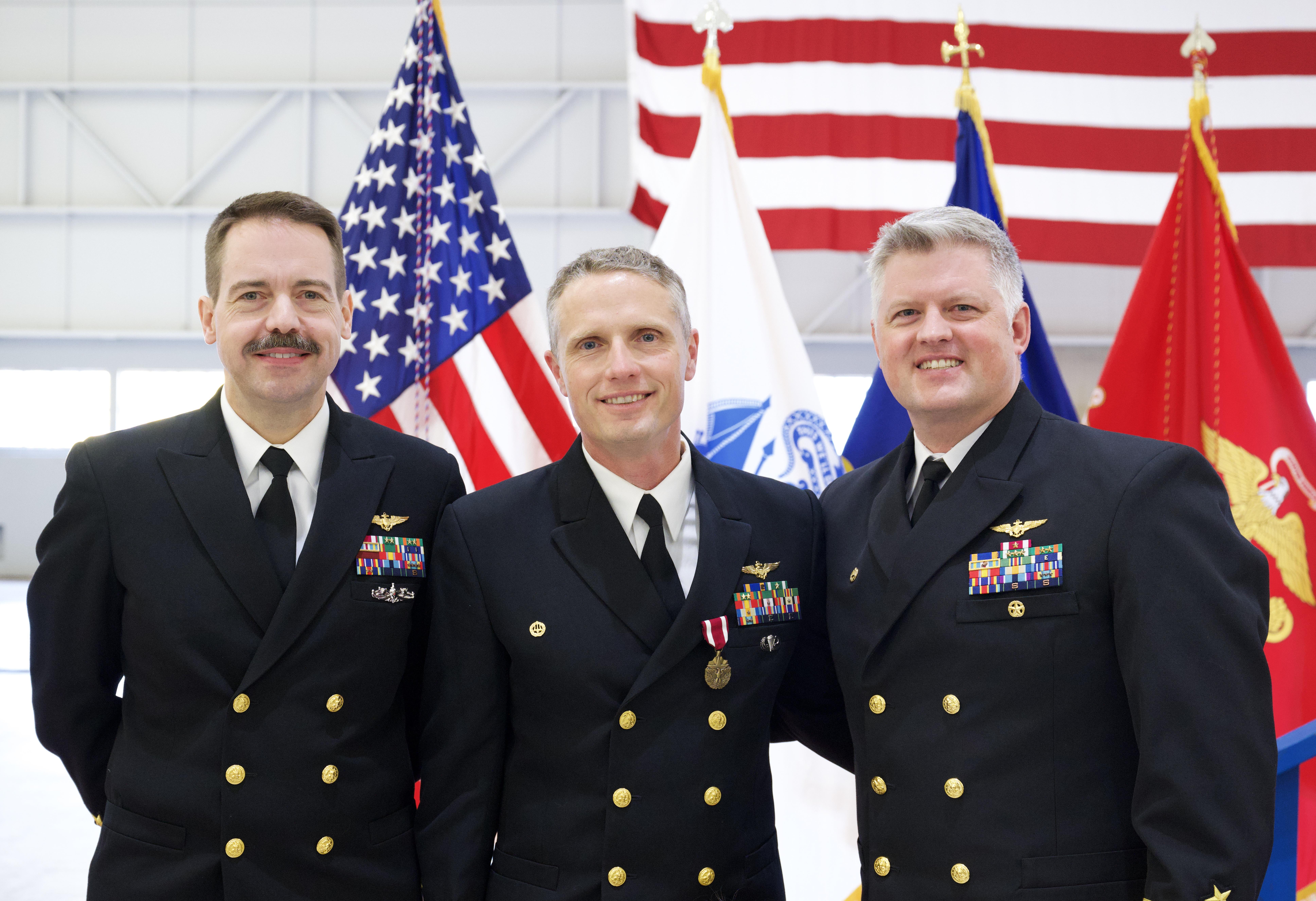 Three naval officers gather for a photo in front of flags inside a hangar space.