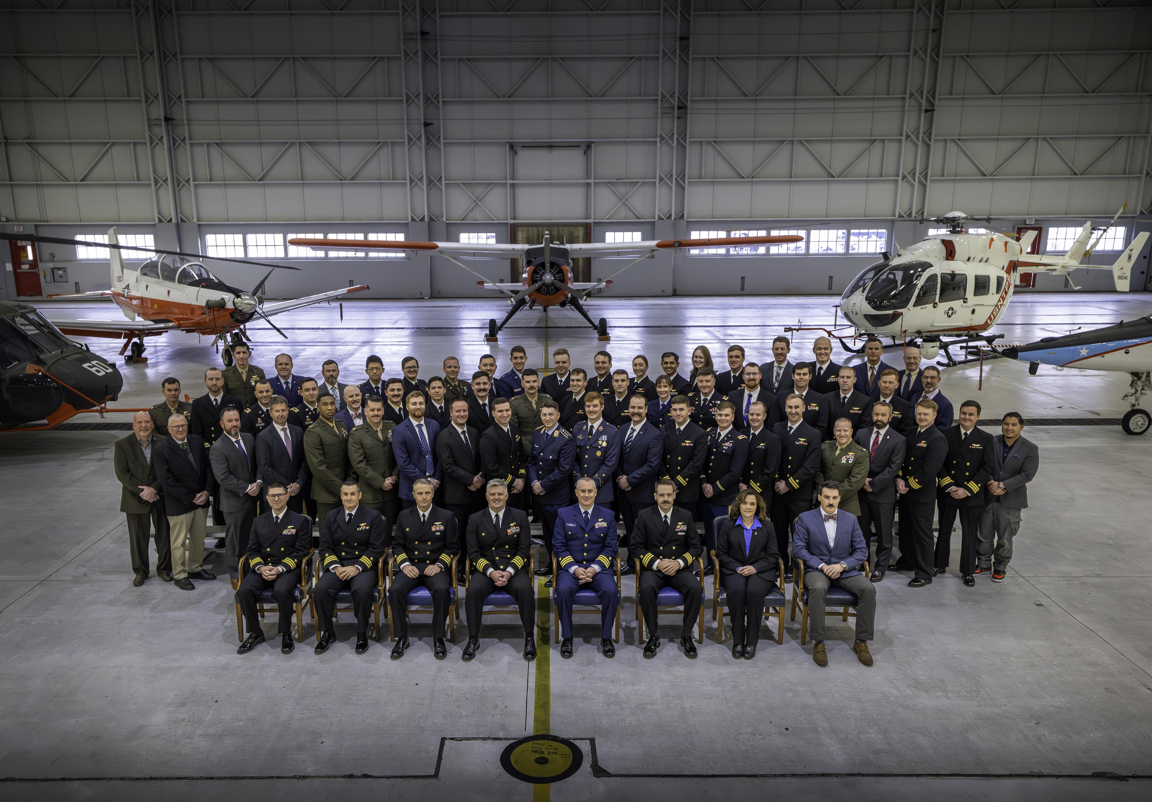 Four rows of people pose for a picture in front of five aircraft. The first row is seated, the back three rows are standing. Most are wearing their military dress uniform. There are a few civilians in business attire.