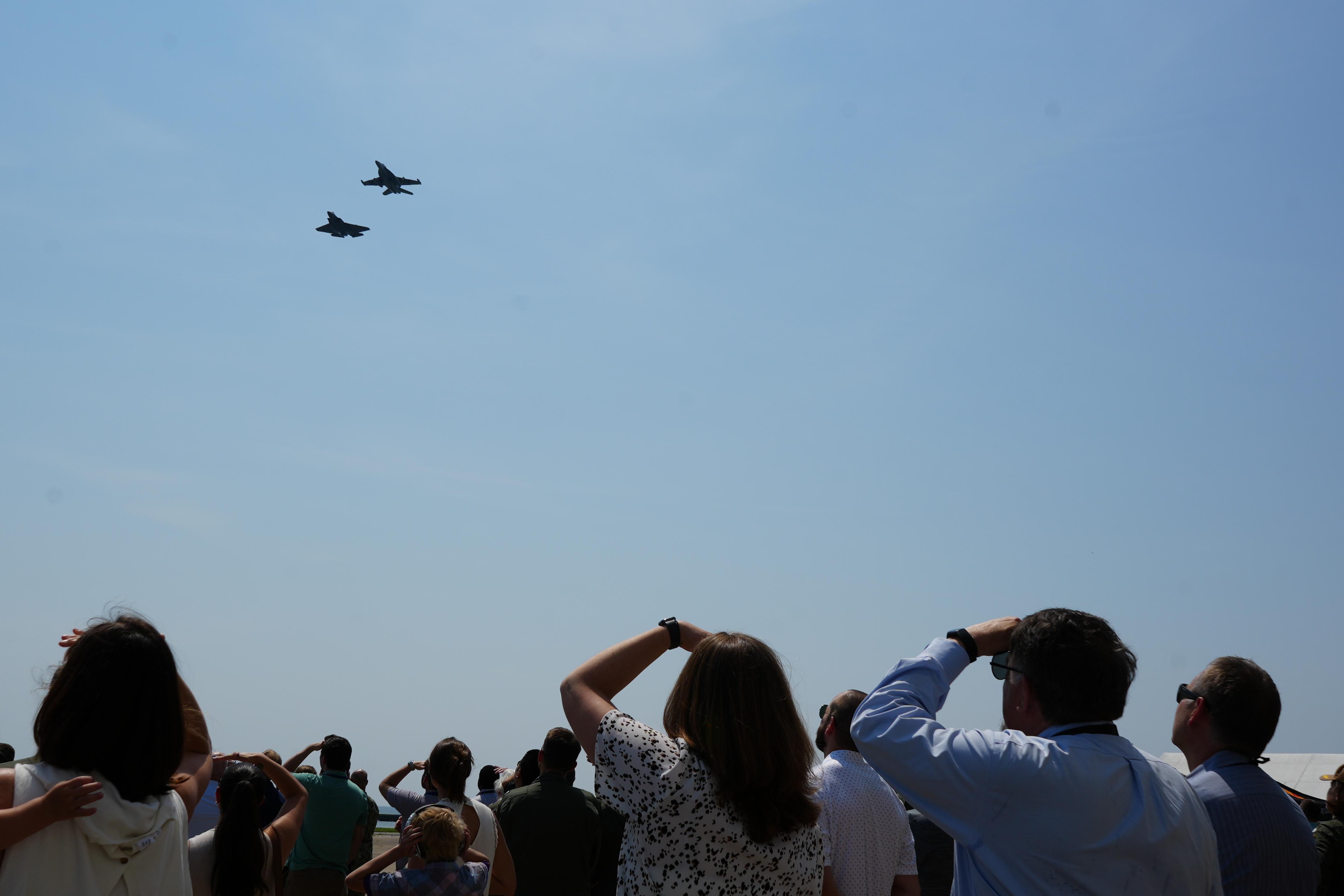 Two jets fly against clear blue skies while change of command ceremony attendees watch from the flightline. 
