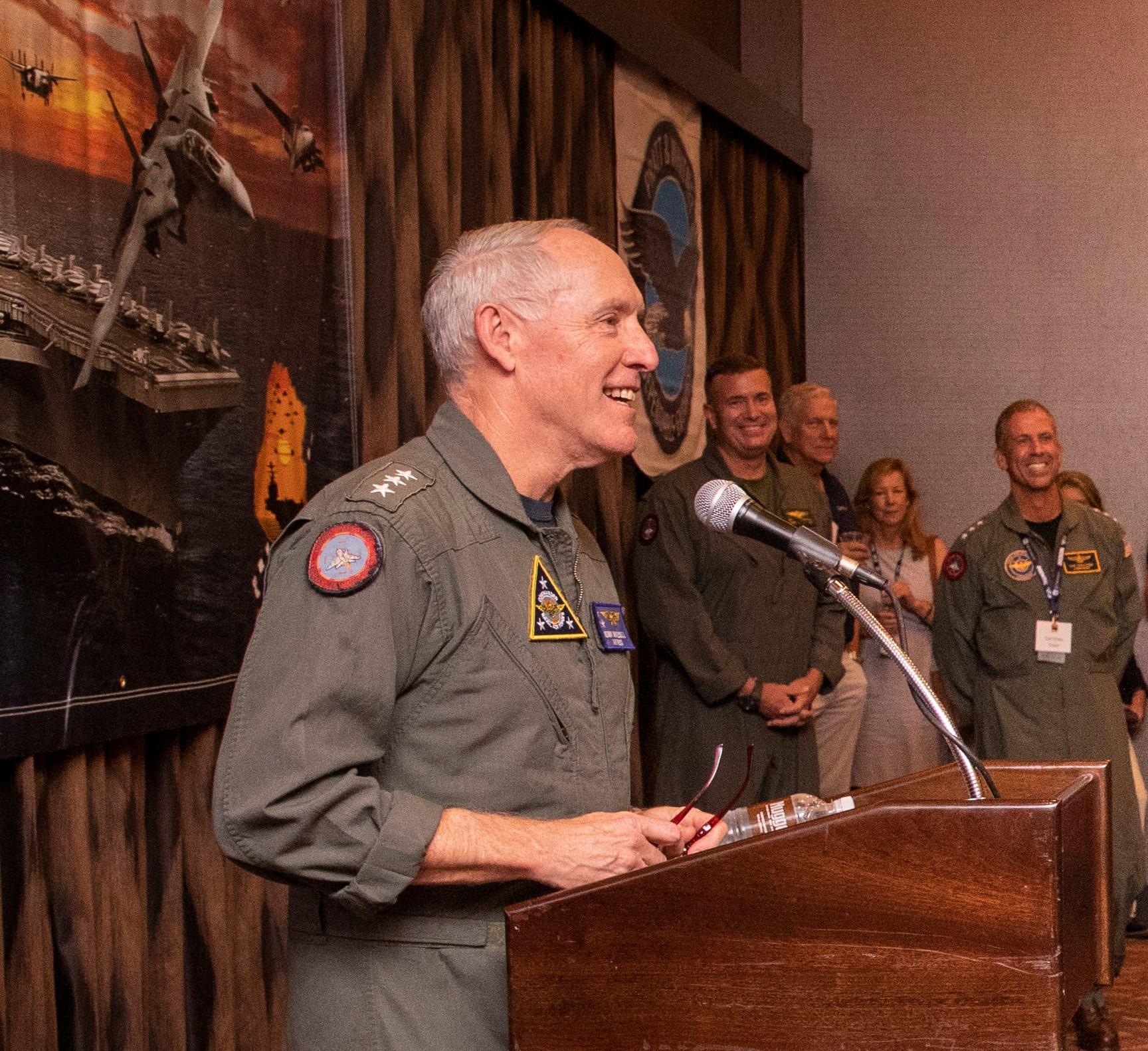 Man in flight suit speaks at podium. 