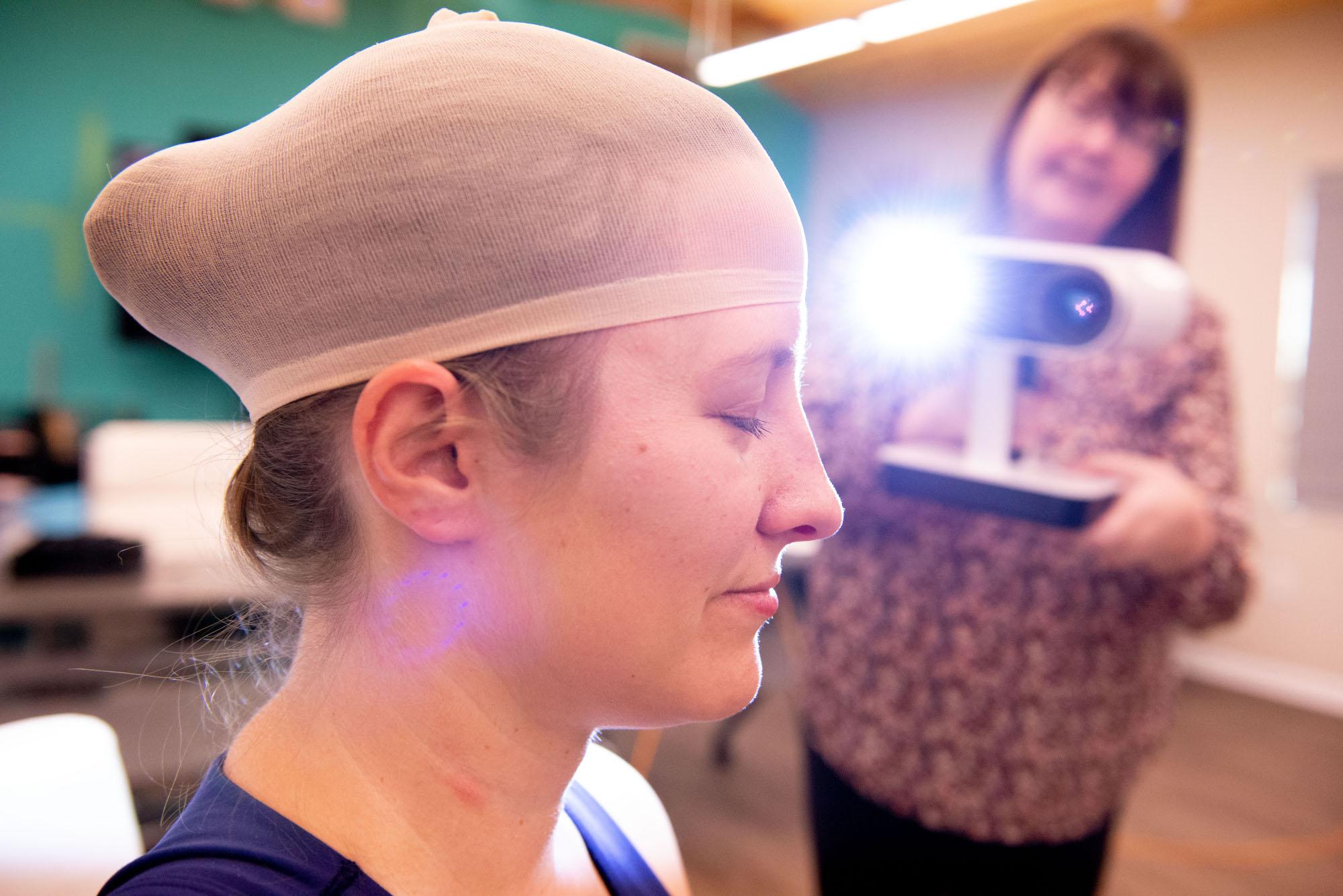 A woman wearing a hair cap sits for head measurements taken using 3D scanning technology.