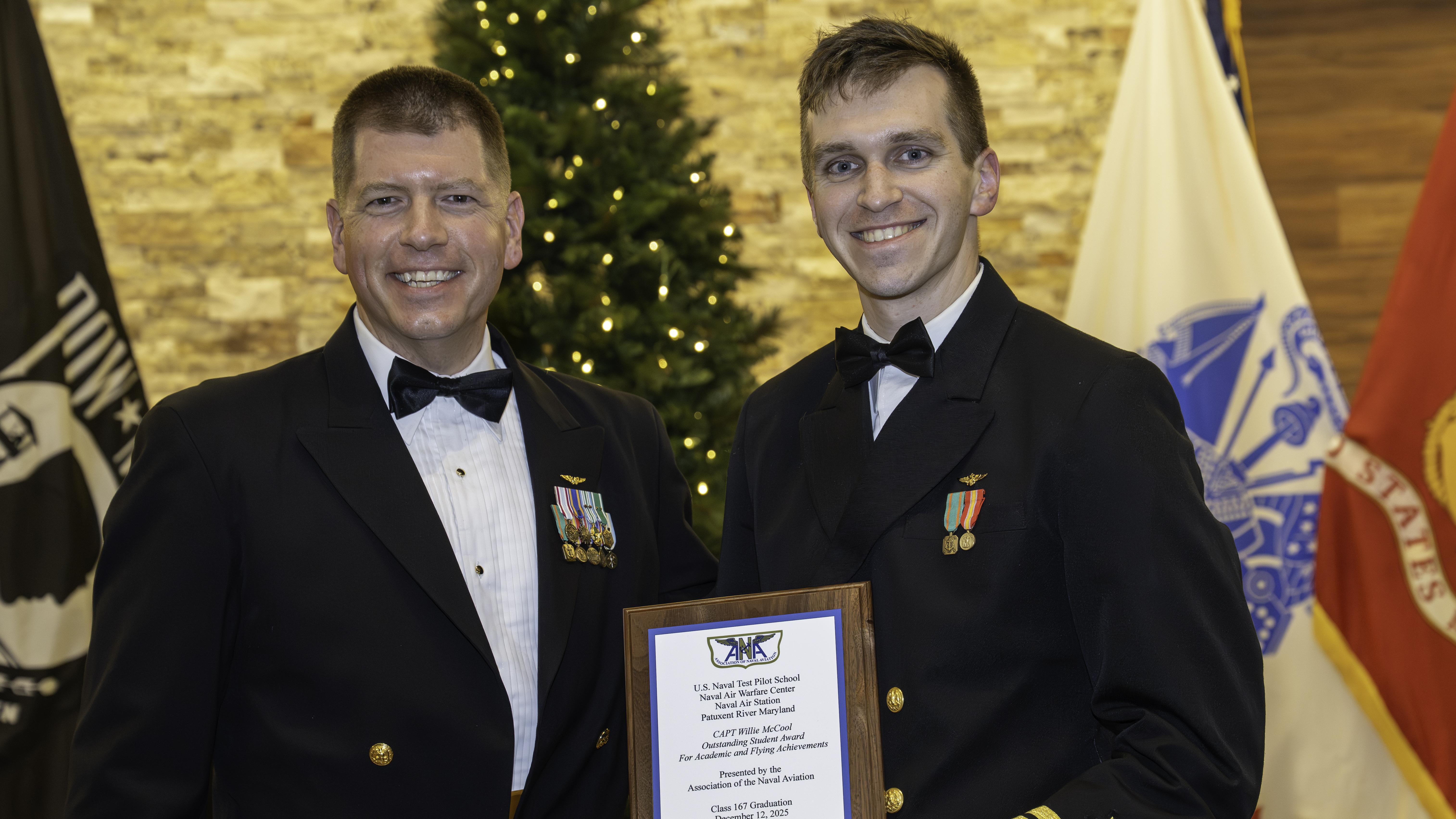 Two Naval Officers pose in front of flags. The one on the left is holding a plaque.