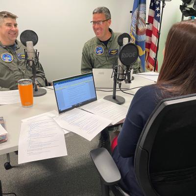 Two pilots in flight suits sitting at a table with microphones recording a podcast.