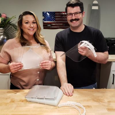 Lt. Gavin “Flanders” Kurey, right, and his wife Katie show some of the hundreds of acrylic face shields they have cut at their metalworking shop for use in making face protection to combat the spread of COVID-19. (U.S. Navy photo by Lt. Gavin Kurey)