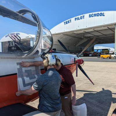 Engineers apply a fiducial tag along the side of a T-6 Texan aircraft in front of a hangar at the U.S. Naval Test Pilot School.