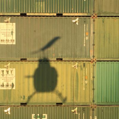 The shadow of a U.S. Naval Test Pilot School (USNTPS) OH-58C Kiowa falls on the face of the Elevated Fixed Platform, a test platform constructed from over 100 shipping containers stacked to form a 38-foot-tall box and capped with steel plates, which USNTPS test pilots recently used to evaluate the hover performance of helicopters near obstacles and inside confined areas.
