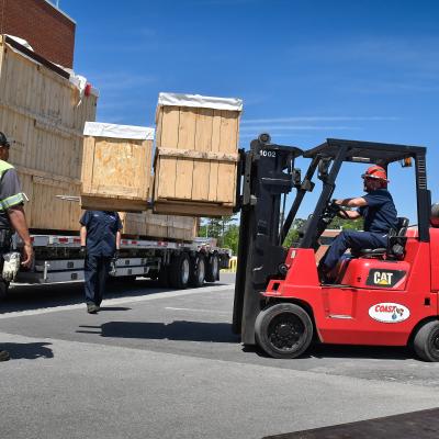Rigging technicians unload a coordinate measuring machine (CMM) from a tractor trailer at Fleet Readiness Center East (FRCE). A CMM is a device that measures the geometry of physical objects by sensing points on the surface of the object with a probe. This machine is one of two new CMMs FRCE acquired capable of large-volume measurement. Depot officials say these additional machines will improve efficiency and reduce wait times caused by maintenance and equipment calibration.