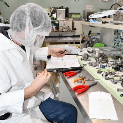 Man in lab coat and hair net and beard cover assembles fuel control unit at clean room work bench