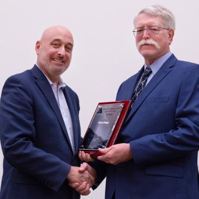 Two men in blue suits shake hands and pose for photo with award plaque between them