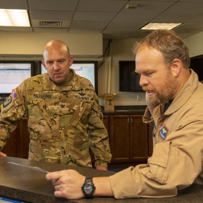 A man in Navy uniform shows a log book to a man in Army uniform in an office space.