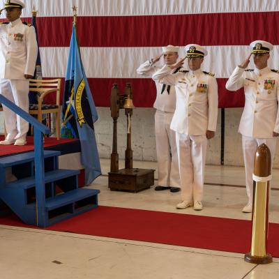Officers in white Navy dress uniforms salute in front of a ceremonial stage.