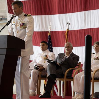 An officer in a white Navy dress uniform speaks behind a podium against an American flag backdrop.
